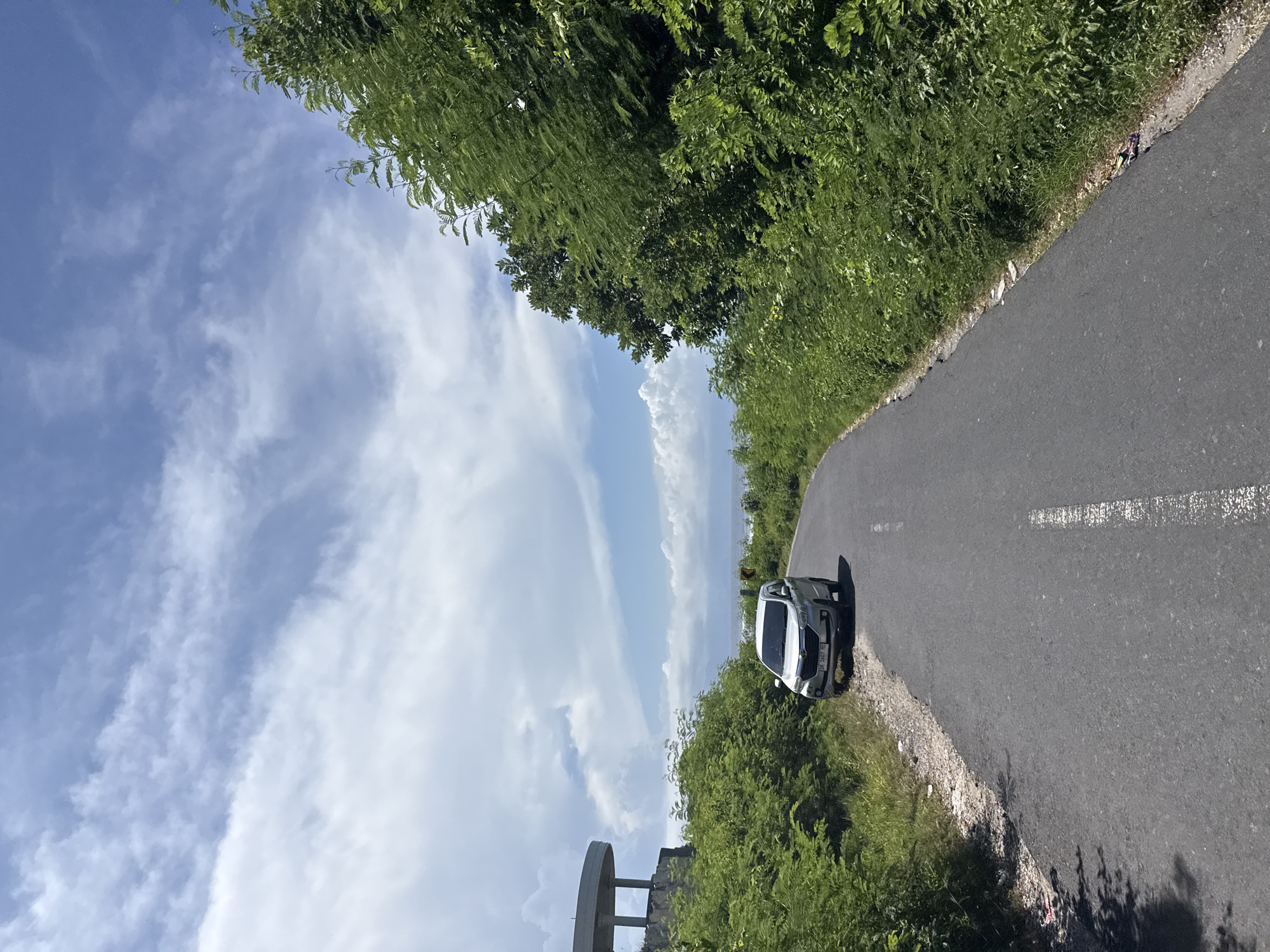 A slightly rotated eye-level shot captures a paved road lined with dense green vegetation under a sky with scattered clouds. A white car is parked on the shoulder of the road. The license plate, "DK 1607 HQ" is visible on the car. A structure that seems to be a partially visible building or pillar is at the bottom left corner. The location is Batununggal, Indonesia.