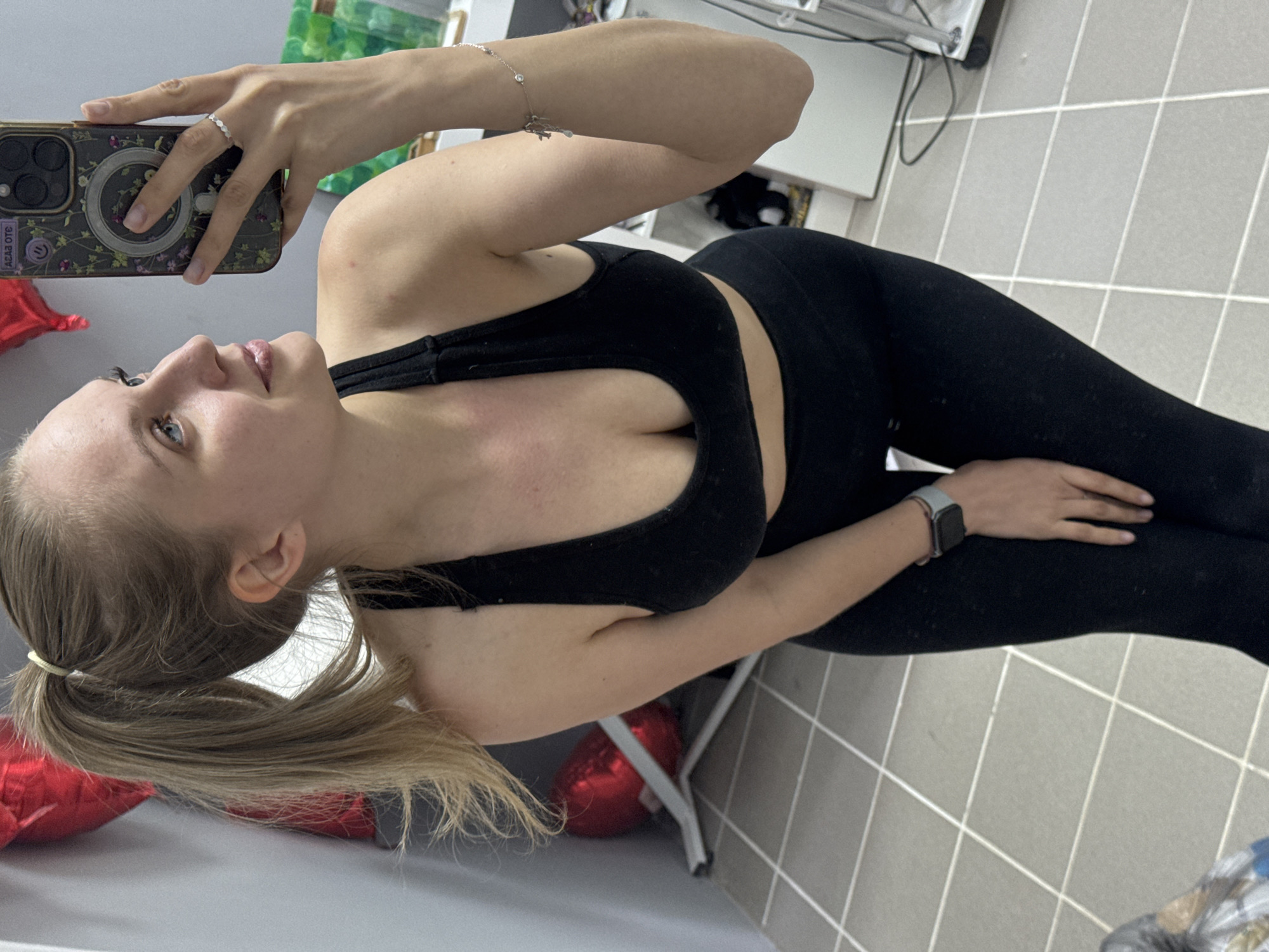 A young woman is depicted in a selfie, with the image rotated 90 degrees counter-clockwise. She has light skin, blonde or light brown hair pulled back into a low ponytail, and blue eyes, looking upwards and slightly to her right with a gentle smile.
She is wearing a black sports bra and black leggings, suggesting athletic or casual attire. In her left hand, she holds a smartphone, which has a distinct patterned phone case, possibly floral. A simple white ring is visible on her left ring finger, and a thin silver bracelet with small charms, including one that looks like an airplane, is on her left wrist. Her right wrist features a smartwatch with a grey/blue band and a dark watch face.
The setting is an indoor room, characterized by light grey square floor tiles with white grout lines. Elements of white furniture, such as a table or cabinet, are visible in the background, alongside some indistinct items. A shiny red object, possibly a foil balloon, is partially visible on the floor on the left side of the frame, next to a metal chair leg.
The primary activity is the woman taking a mirror selfie. On the phone case, some reversed text is visible, appearing to be "ACADEMY" or a similar word as part of a design. The image captures an indoor scene without specific time-of-day indicators. The location context for this image is Izhevsk, Russia.