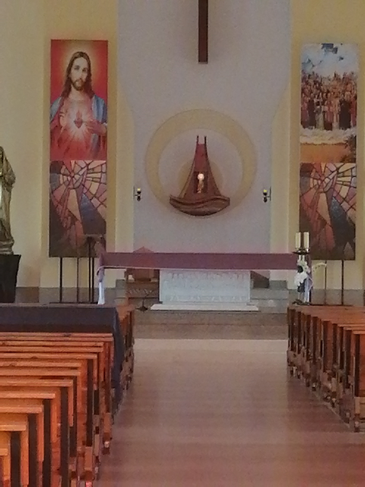 The image depicts the interior of a church or chapel, with a focus on the altar area and pews. On the left wall hangs a banner with an image of Jesus with his hands outstretched, showing his heart, set against a red background. Below this is a stained-glass-style panel with geometric patterns. On the right wall, a similar stained-glass-style panel is visible, and above it is a banner depicting a crowd of people. In the center, behind a maroon cloth draped over a structure, is a white altar or communion table. Above the altar is a circular motif with a boat-like sculpture hanging within it. A statue of a religious figure is partially visible on the far left, standing in a dark planter. Rows of wooden pews are arranged in the foreground, leading towards the altar. The walls are painted a light color. The overall scene suggests a place of worship. There are no individuals clearly visible in the image performing any actions. The lighting is soft, suggesting indoor lighting. This scene is not located in Lạc Dương, Vietnam.