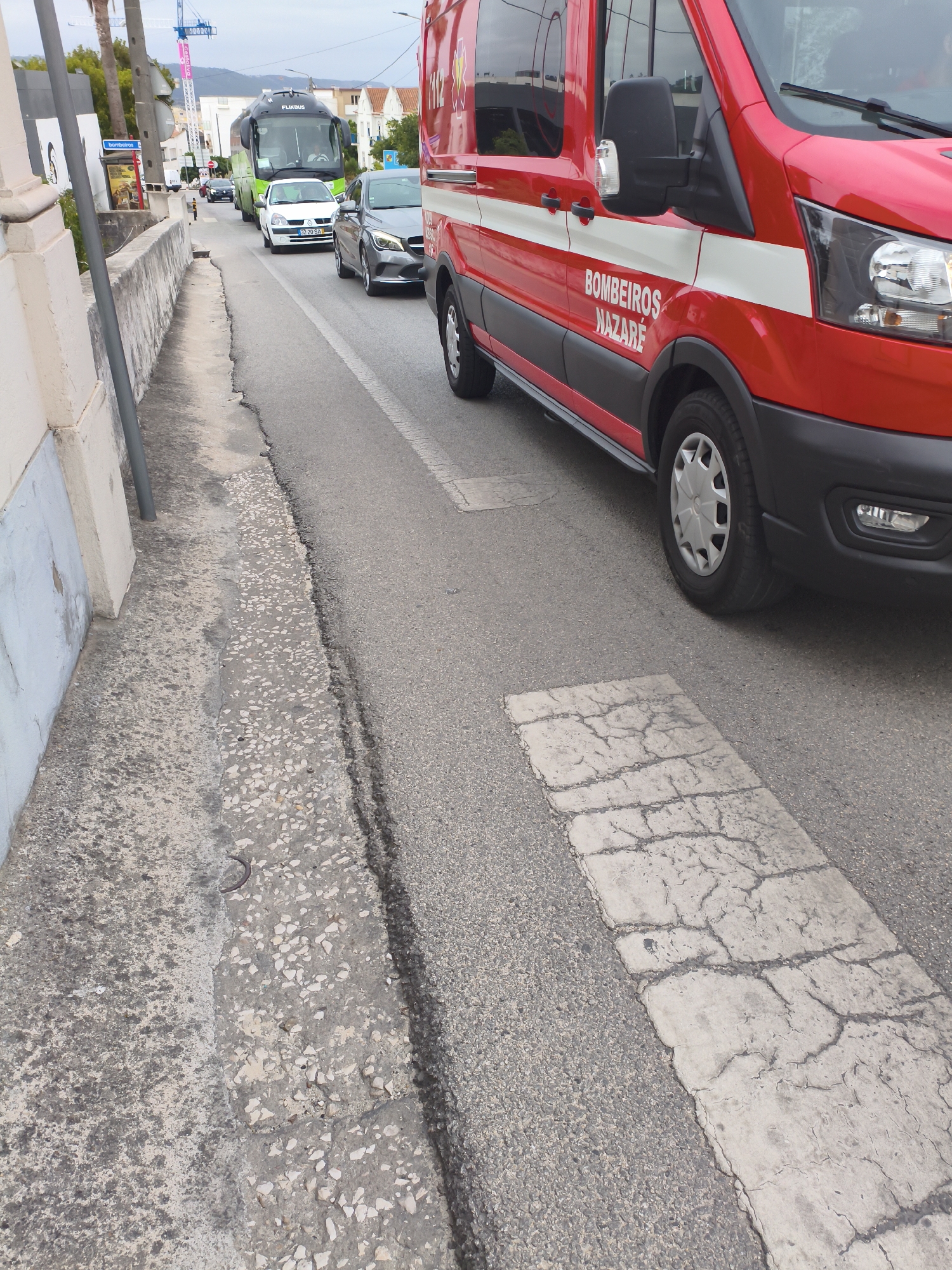 The image shows a street scene in Nazaré, Portugal, on an overcast day. A red fire truck with white stripes and the words "BOMBEIROS NAZARÉ" is prominently featured in the foreground, parked or driving on the right side of the road. The truck has a large side mirror and a part of its front grille and headlight are visible. To the left of the fire truck, a grey car is visible in traffic. Further down the road, a white car and a green bus with "FLIXBUS" written on it are also present in traffic. The road appears to be paved with asphalt and has a white textured stripe marking a pedestrian crossing or speed bump. The left side of the image shows a concrete sidewalk with a textured surface and a wall. The overall environment suggests a town or urban setting. There are no people clearly visible in the image.