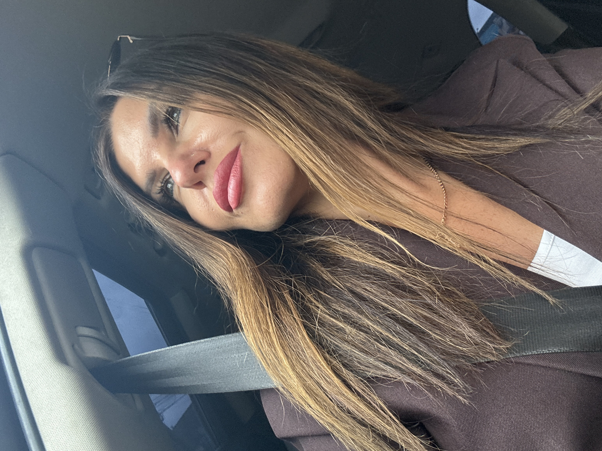 A woman with long, brown, highlighted hair is in the driver's seat of a car. She is looking out the window to the right, with her head tilted upwards. She is wearing a dark brown blazer over a white top and a delicate gold chain necklace. A seatbelt is visible across her chest. The car has a sunroof, and the sky is visible through it. The background is dark and out of focus, suggesting the car is in motion. The location is described as the city of Chaikovsky, Russia.