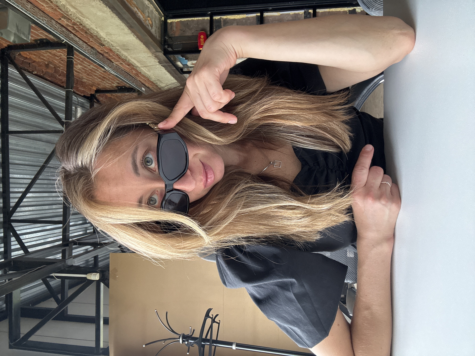 A young woman with blonde hair and sunglasses is sitting at a table, looking at the camera. She is wearing a black top and a silver necklace. She is leaning on the table and her hand is resting on her chin. The background is a brick wall and some industrial-looking metal structures. The scene appears to be indoors, possibly in a cafe or restaurant. There is no indication in the image that suggests the location is the city of Chaikovsky, Russia.