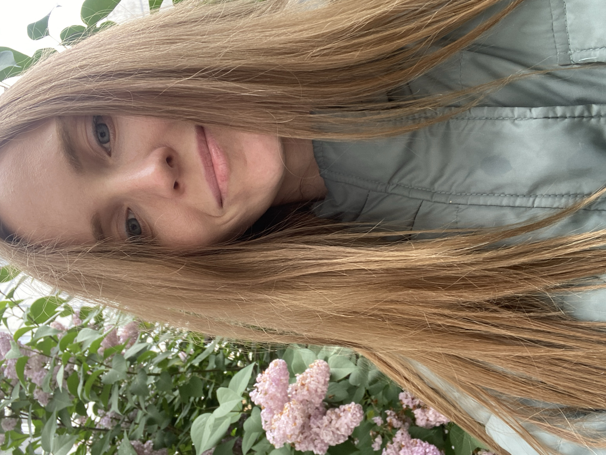 A Caucasian woman with long, blonde hair smiles and looks at the camera. She is wearing a light green jacket. Behind her, there is a bush with pink flowers and green leaves. The image appears to be taken outdoors, possibly in a park or garden.