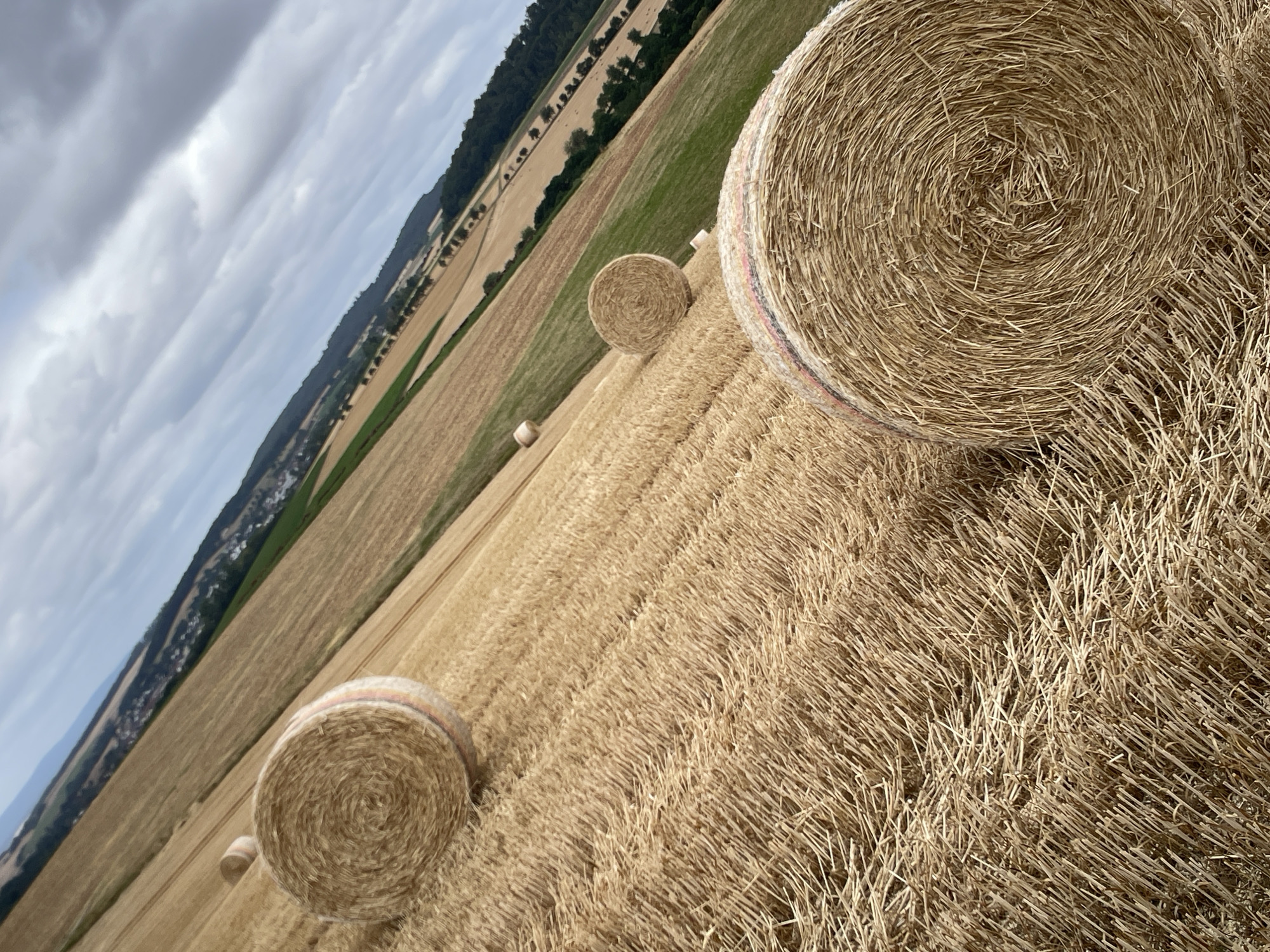 This is a tilted, close-up, eye-level, outdoor, daytime, medium shot of a hayfield in Asterode, Germany. Several round bales of hay, mostly brown and tan, are scattered across the field. The bales are arranged at different angles and distances from the camera. The field is covered in dry, cut wheat stalks. In the background, there are green fields and trees, with a village and hills beyond. The sky is overcast with dramatic clouds.