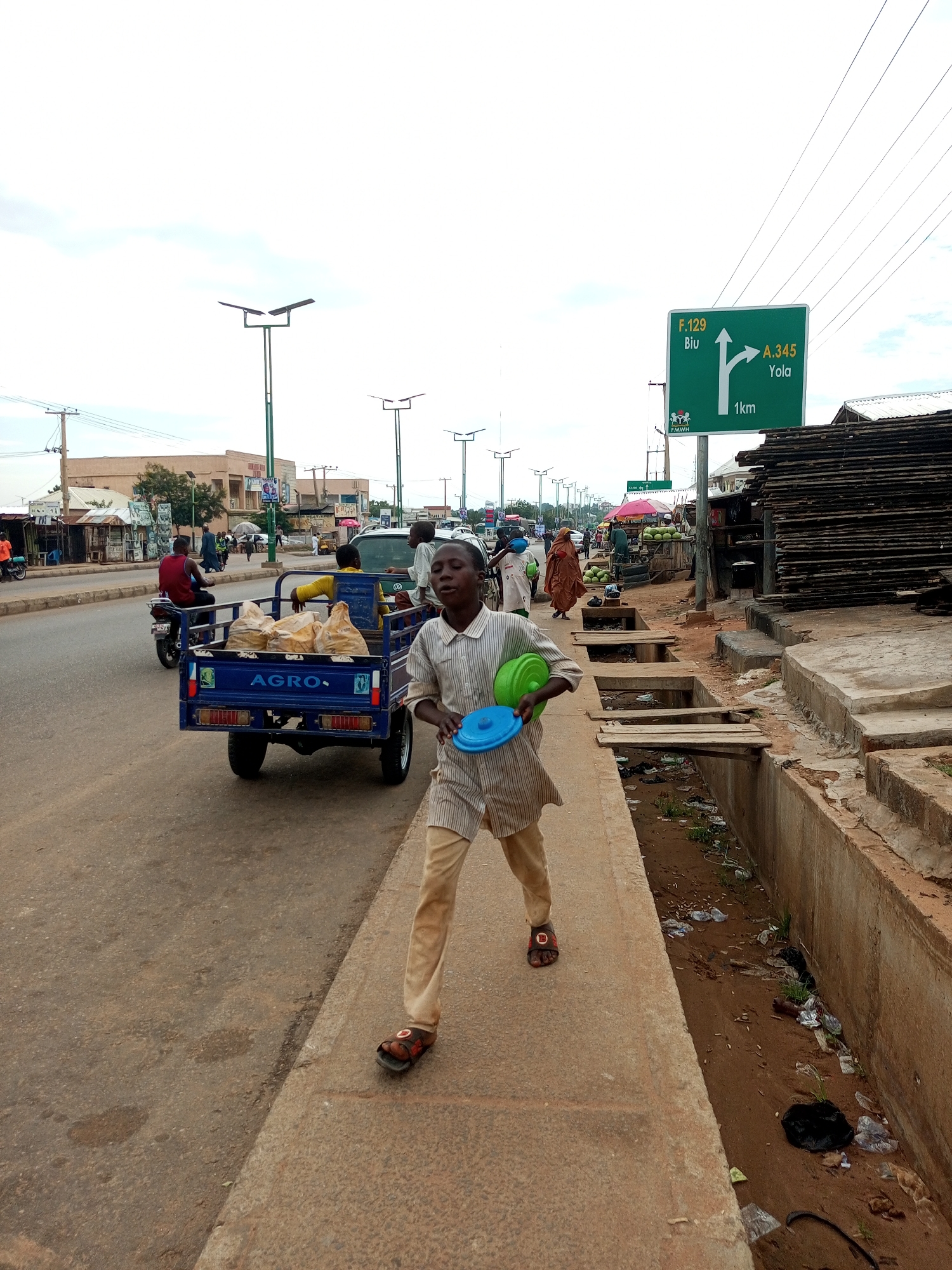 The image depicts a street scene in Gombe, Nigeria, likely along a main thoroughfare. In the foreground, a young boy wearing a striped shirt and light-colored trousers walks on a concrete sidewalk, carrying two plastic bowls, one blue and one green. He is facing slightly away from the camera. To his left, a blue trailer pulled by a motorcycle is loaded with several large bags, possibly containing goods. A man in a red shirt is seated on the motorcycle. In the background, several vehicles, including a car and other motorcycles, are visible on the road. Buildings line the street, some appearing to be shops or market stalls. There are also solar-powered streetlights and a green road sign indicating directions to "F.129 Biu" and "A.345 Yola" with a distance of "1km." The environment appears somewhat dusty, and a drainage ditch with visible debris runs alongside the sidewalk. The sky is overcast.