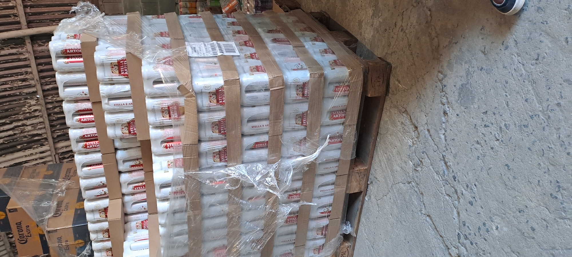 The image shows a large stack of Stella Artois beer cans, shrink-wrapped and secured to a wooden pallet. The pallet is resting on a concrete floor, with a corrugated metal wall or structure in the background. To the left of the pallet, stacks of Corona Extra beer boxes are visible, also wrapped in plastic. One person's foot, wearing a sneaker, is visible in the upper right corner, suggesting the photo was taken by someone standing near the pallet. The lighting appears to be natural, suggesting it might be daytime, though the indoor setting makes it difficult to determine the exact time of day. The overall impression is one of a storage or warehouse environment where beverages are being kept. The visible text includes "Stella Artois" and "Corona Extra," indicating the brands of beer.
