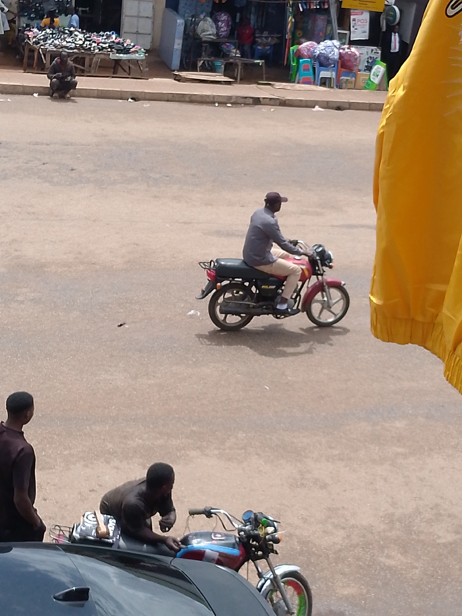 In Bukuru, Nigeria, a street scene unfolds with multiple individuals and vehicles. In the foreground, a dark-colored car is partially visible, with two men standing nearby. One man, on the left, faces away from the camera, wearing a dark short-sleeved shirt. The second man, on the right, leans over a motorcycle, appearing to adjust something. This motorcycle has a black seat with a bag on it, and its front is adorned with colorful decorations, including green and red elements.
Further into the scene, a man wearing a light gray long-sleeved shirt and beige pants rides a red motorcycle. He has a dark baseball cap on his head and is looking forward. Behind him, a stall displays a large quantity of footwear. A person in a dark shirt is crouched down in front of this stall. In the background, various market stalls and structures are visible, with some people milling about. The ground is paved with a light-colored, sandy material. A large yellow fabric, possibly clothing or a banner, is draped on the right side of the frame, obscuring part of the background.