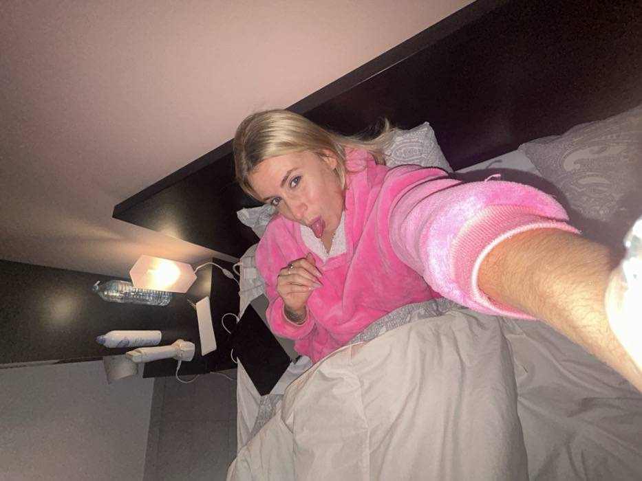 A blonde woman in a pink robe lies in bed, sticking her tongue out at the camera. Her head is tilted to the side, and her arm is outstretched, likely holding a phone for a selfie. A headboard is visible behind her, along with pillows. A lamp and several other objects are on a shelf beside the bed. The room is dimly lit.