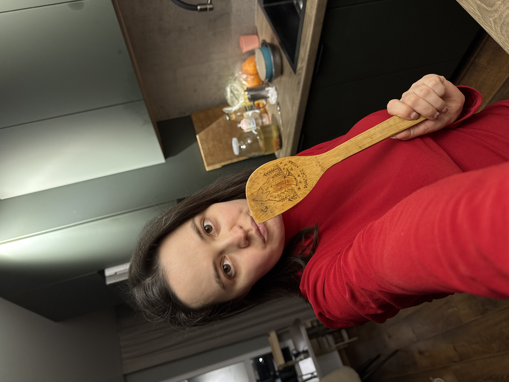 A woman in a red long-sleeved shirt is holding a wooden spoon with carvings on it, posing for a selfie. The photo appears to be taken indoors in a kitchen setting. A countertop with a sink, some kitchen utensils, and food items are visible in the background. The photograph does not contain any information to establish that it was taken in Izhevsk, Russia.