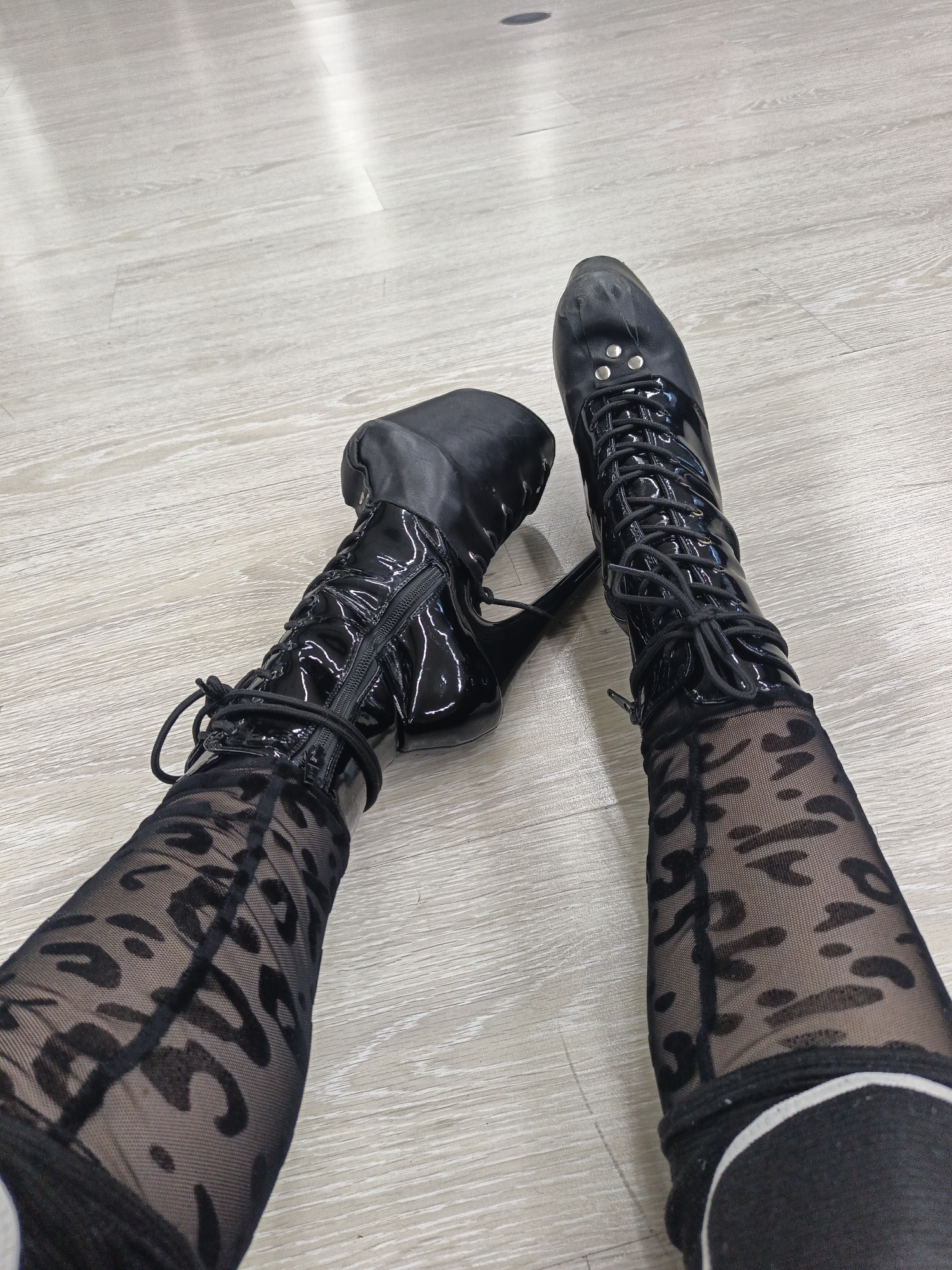 The image presents a first-person, overhead view of a person's legs and feet, showcasing their footwear and hosiery. The subject is wearing black patent leather, lace-up, high-heeled boots and sheer black tights with a distinct dark pattern, possibly leopard print or an abstract design.
Both boots feature extensive lacing up the front and zippers along the inner ankle. The left boot clearly reveals its high heel and a small platform, characteristic of high-fashion or performance footwear. The right boot's toe area appears worn and scuffed, indicating prior use. The glossy surface of the patent leather reflects overhead lighting, as do subtle areas of the light-colored floor.
The floor beneath the subject's legs is light-toned, appearing to be wood laminate or a similar material with a faint grain texture. The setting is indoors, under bright artificial lighting. The activity captured is static, likely a pose to display the outfit, with the person either seated or lying down with their legs extended. There are no discernible emotional expressions, visible text, specific location identifiers, or indications of outdoor weather or time of day. The location remains unknown.