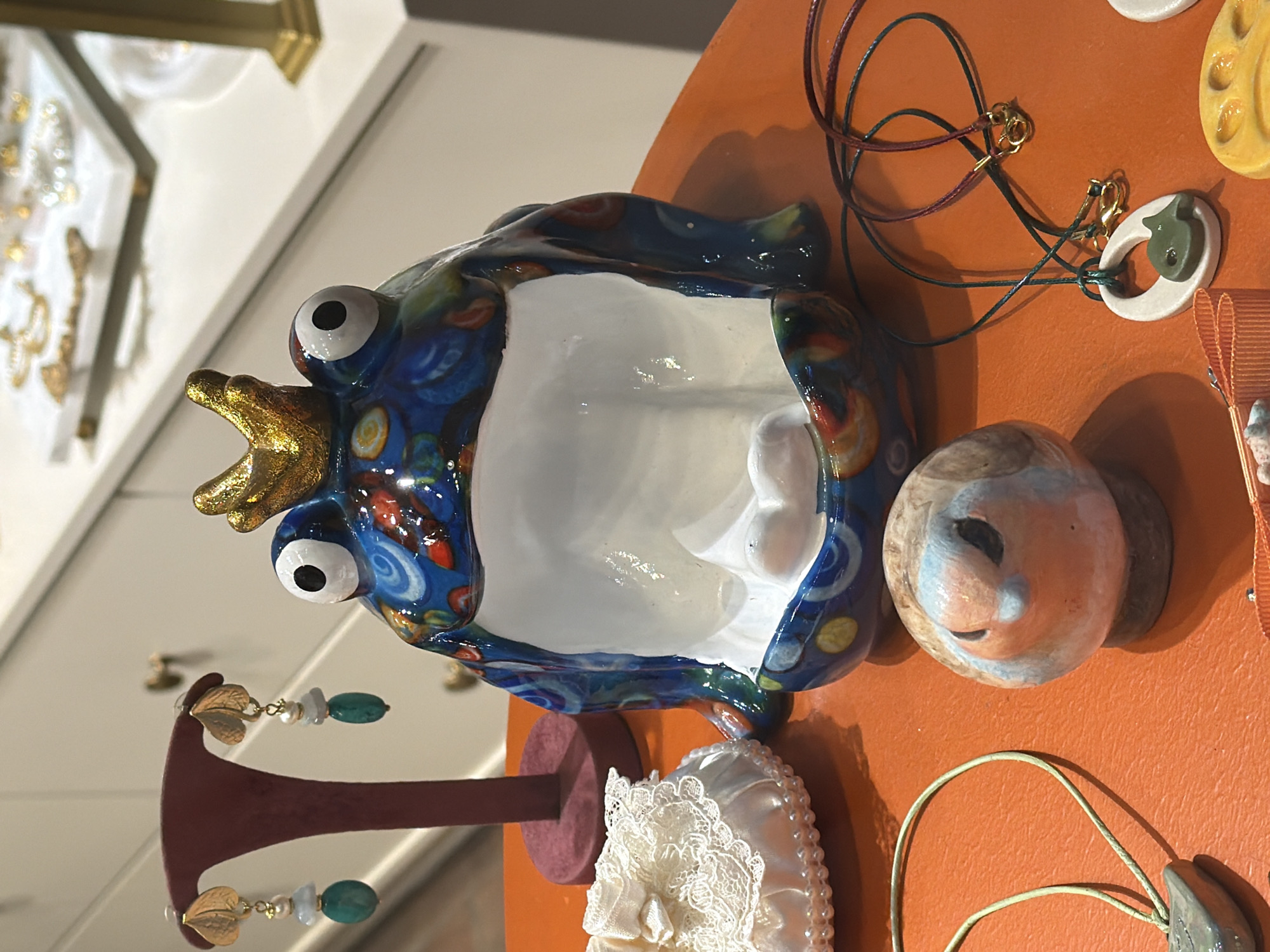 A close-up, eye-level shot shows a collection of decorative items arranged on an orange surface, possibly a table or shelf. The background is blurred, but appears to be a retail display with white cabinetry and shelves.
In the foreground, a ceramic figurine of a frog wearing a golden crown is the central subject. The frog is predominantly blue with colorful patterns of red, yellow, and white. Its eyes are large and white with black pupils, and its mouth is open, revealing a white interior. To the left of the frog is a small, white, lace-covered drawstring pouch with pearl-like embellishments around its rim.
To the left of the frog, on a dark red stand, hang a pair of earrings. Each earring features a gold leaf charm from which dangle two beads: one pale blue and one darker teal.
In the lower part of the image, a small, round ceramic bird figurine with an orange breast and blue and grey plumage is positioned to the right of the frog. In front of the bird are two necklaces. One is a green cord with a white oval pendant that has a small, dark green fish charm. The second necklace has a darker cord and a gold clasp. Further to the right, a yellow ceramic item with a paw print design is partially visible.
The scene is brightly lit, with a soft focus in the background that highlights the colorful objects in the foreground. The overall impression is of a display of artisanal or handcrafted jewelry and decorative pieces. The image does not contain any identifiable individuals, actions, or interactions. The location in Üsküdar, Turkey is not visually discernible from the provided image.