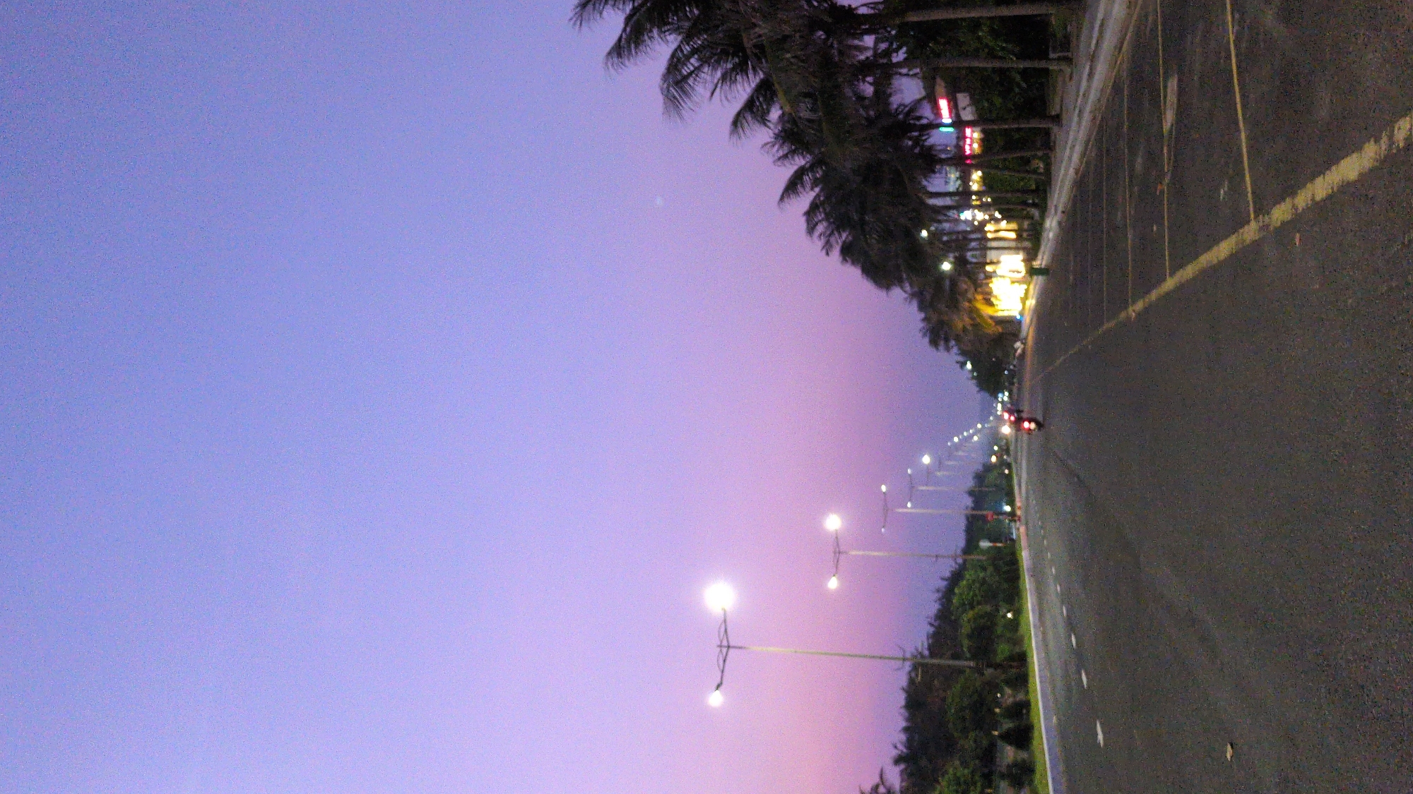 The image captures a street scene in Tuy Hòa, Vietnam, at twilight. The sky is a gradient of purple hues. Palm trees line the left side of the frame, bordering the road. Streetlights illuminate the road, stretching into the distance. A motorcycle with red taillights is visible further down the road. The road itself is dark asphalt with painted yellow lines. Buildings and greenery are present along the roadside.