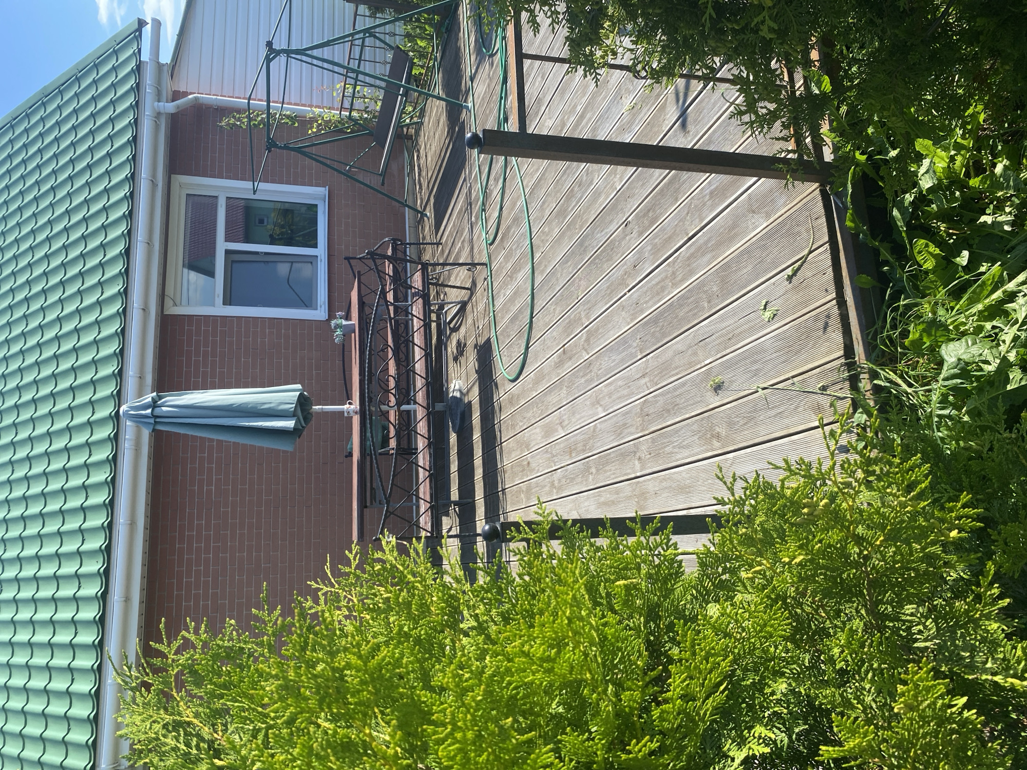 This is a high-angle, full shot of a wooden deck in front of a brick house in Bol'shoe Zaborod'e, Russia. The deck is empty except for a green garden hose coiled on the ground. To the left, a closed teal umbrella stands on a patio table with two benches, a small potted plant, and a cup. Behind the table and benches, a green metal swing set is visible. A large green evergreen tree is positioned in the foreground on the left, and another smaller one is on the right, partially obscuring the view of the deck and the house. The house has a green metal roof, a white gutter, and a single window with a white frame. The sky is blue with some clouds.