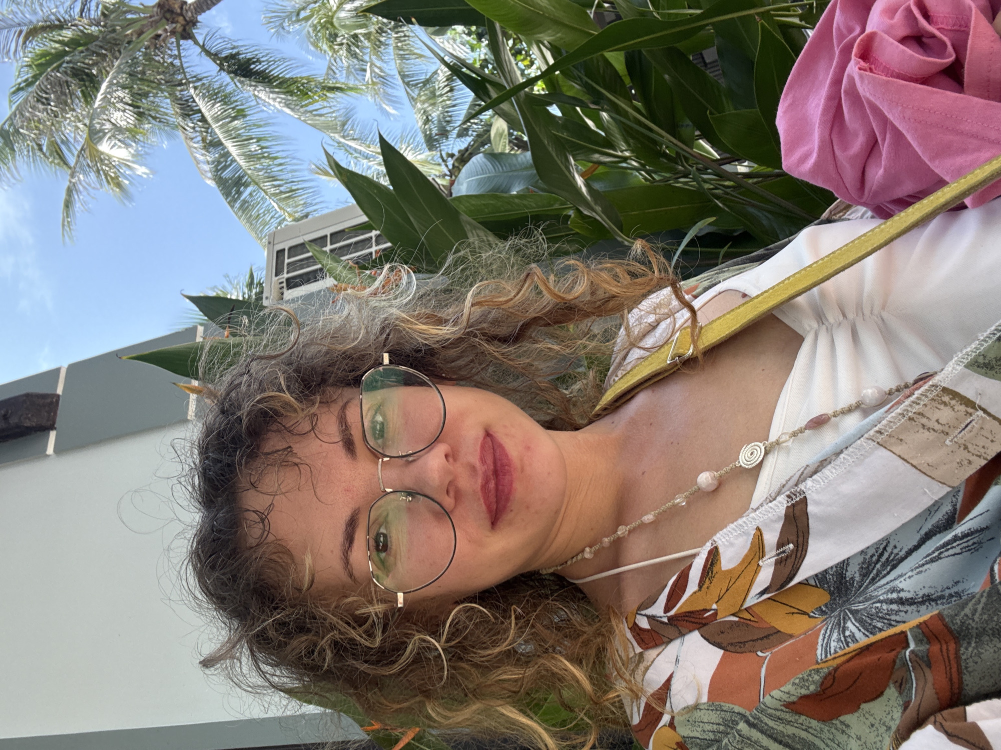 A woman with curly blonde hair and glasses is captured in a close-up, outdoor shot. She is wearing a white top layered with a patterned blouse featuring tropical leaves and flowers in shades of brown, orange, green, and white. A long beaded necklace with pink and clear stones adorns her neck. A bright yellow strap, likely from a bag, crosses her chest. Partially visible in the background are lush green tropical plants, palm trees, and a clear blue sky, suggesting a warm, sunny climate. The setting appears to be a resort or a location with abundant greenery, consistent with the coastal resort town of Mũi Né, Vietnam, known for its beaches and palm-lined shores. A crumpled pink garment rests on her right side.