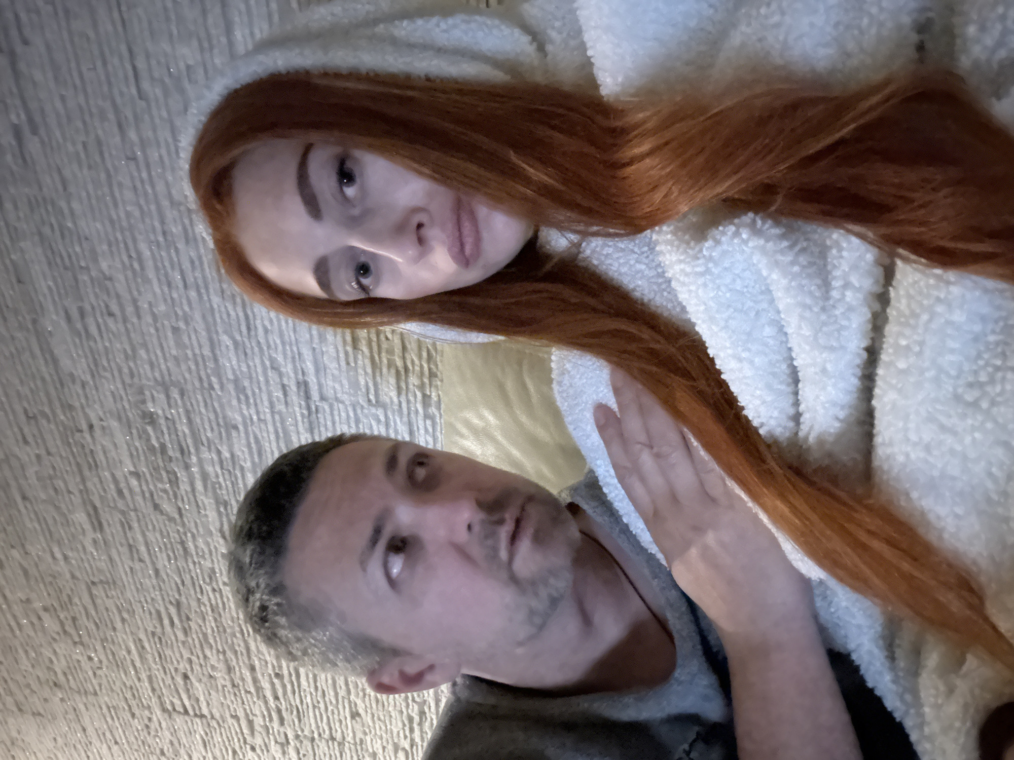 This image captures two individuals, a woman and a man, in a relaxed indoor setting. The woman, positioned closer to the viewer on the left, has long, flowing reddish-brown hair and fair skin. She is wearing a cream or white fluffy, hooded garment and looks directly at the camera with a neutral, slightly pensive expression. Her arm is visible, resting gently on or near the man.
The man is situated slightly behind and to the right of the woman. He has short, greying hair and stubble, and is wearing a grey top. His gaze is directed upwards and to the right, away from the camera, also with a neutral expression. Both individuals appear to be lying down or reclining, suggesting a moment of rest or intimacy.
The background consists of a light-colored, textured wall, possibly made of stucco or a similar material, creating a subtle, vertical pattern. The lighting is soft and appears to be artificial, indicative of an indoor environment, making the specific time of day difficult to determine but suggesting evening or night. There is no visible text in the image. The location context provided is Budva, Montenegro.