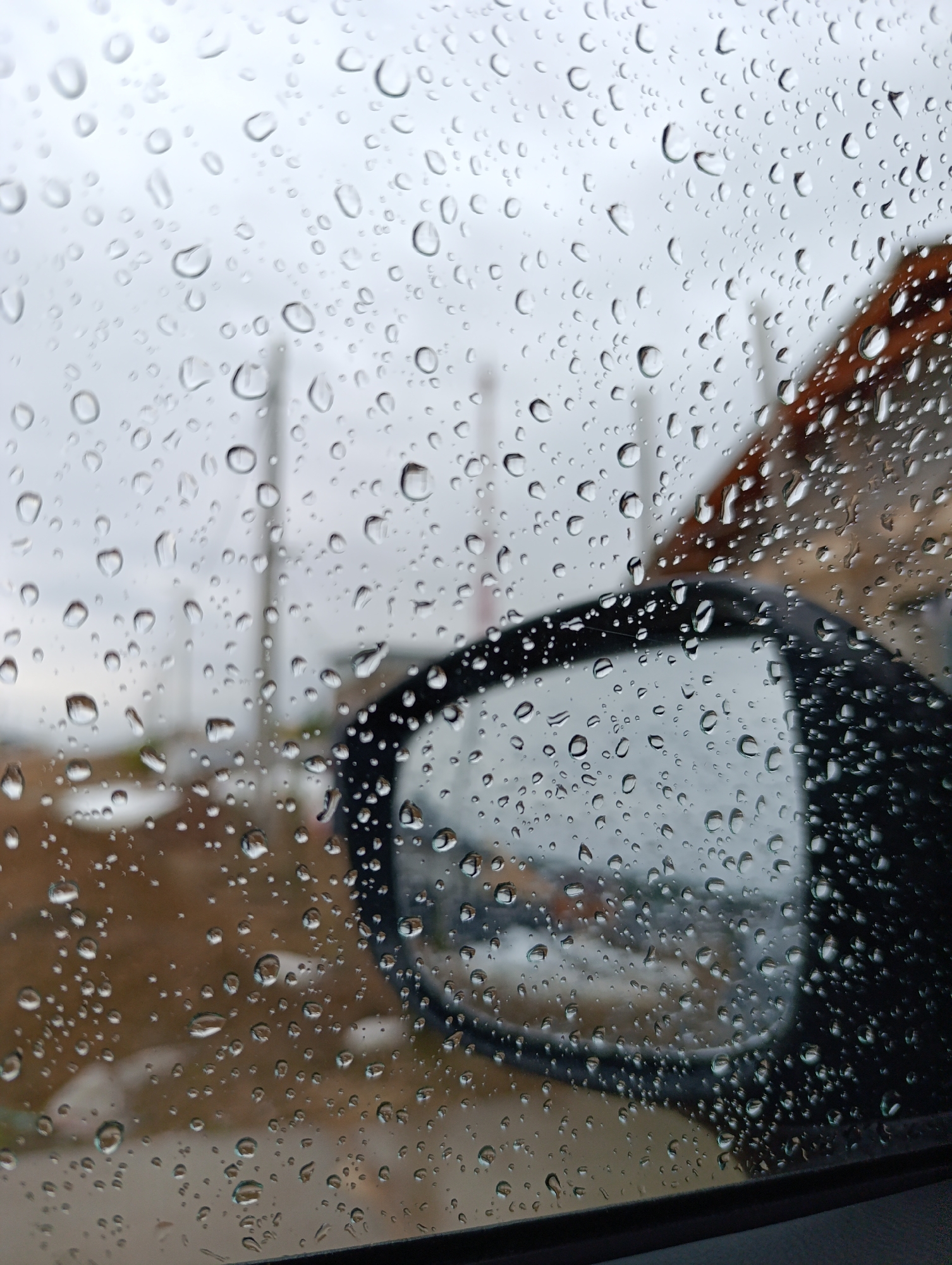 The image shows a rainy view through a car window. Raindrops cover the glass, partially obscuring the outside scene. A black car side mirror is visible on the right, reflecting a blurry image of the surroundings. Outside the car, the landscape appears wet and overcast. Two poles can be seen in the background. In the reflection of the mirror, it seems to be a small town. The visible architecture and layout of the area are consistent with a possible location in Chaikovsky, Russia. The details of the location are unclear, and the location cannot be 100% proven.