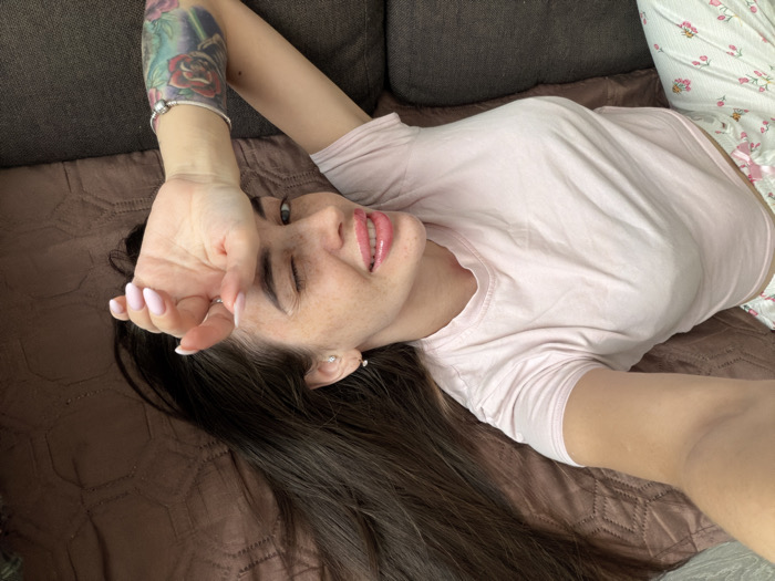 The scene depicts a female individual lying in an indoor residential setting, ostensibly within the city of Novosaratovka, Russia. The individual, an adult with long dark brown hair, is positioned with her head towards the upper left of the frame, looking directly towards the camera. Her face features freckles, and she wears light pink lipstick. Her left eye is partially closed, while her right eye is open. A silver stud earring is visible in her left ear. Her left arm is raised, with her hand partially covering her forehead. The nails on her visible left hand display a pink and white French manicure. A silver charm bracelet is present on her left wrist. A colorful tattoo featuring red roses and green foliage is visible on her left upper arm. She is attired in a light pink short-sleeved t-shirt and white pajama bottoms with small pink and green floral patterns. She is lying on a surface covered by a dark brown, quilted blanket or throw with a diamond pattern. A dark gray sofa cushion is visible behind her head.