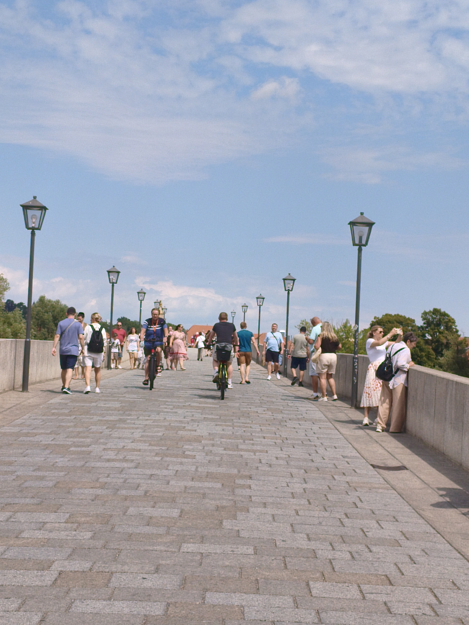 The image depicts a sunny day on the Steinerne Brücke (Stone Bridge) in Regensburg, Germany, a historic stone arch bridge spanning the Danube River. The foreground shows the textured cobblestone pavement of the bridge, worn smooth by time and traffic. Numerous people are walking and cycling across the bridge, enjoying the pleasant weather.
On the left side of the bridge, a man in a blue shirt and dark shorts walks away from the camera, accompanied by a woman in a white top and shorts carrying a backpack. Further down the bridge, a man in a blue t-shirt and shorts cycles towards the camera. On the right side of the bridge, a couple is taking a photo with a smartphone. The woman in a white top and floral skirt is holding the phone, while the man in a light blue shirt and beige shorts looks on.
The bridge is lined with traditional lampposts, casting long shadows on the pavement. In the background, trees and buildings can be seen, suggesting the urban environment of Regensburg. The sky is a clear blue with scattered white clouds. The overall atmosphere is one of a vibrant and lively public space, filled with both locals and tourists.