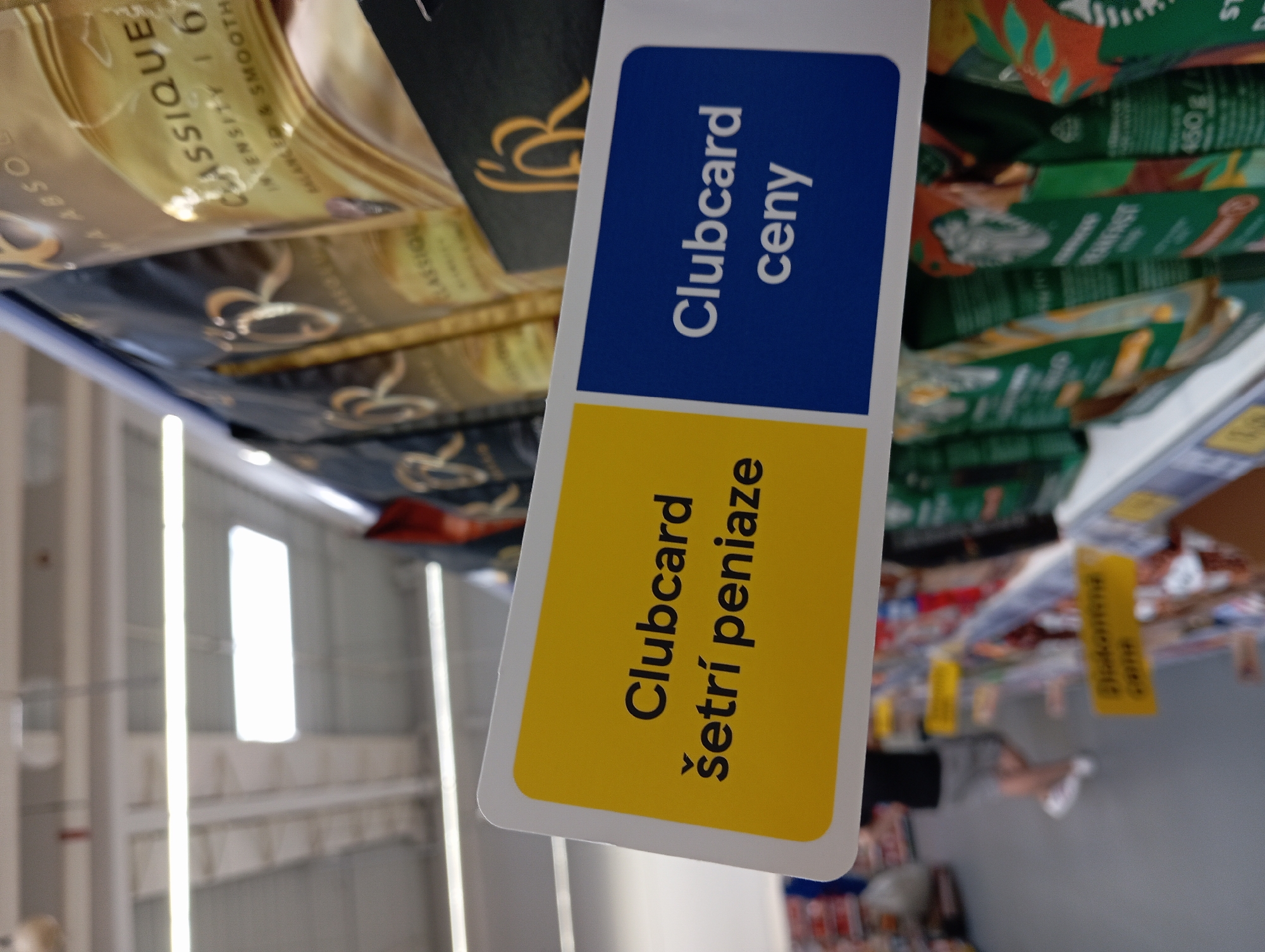 The image shows a close-up of a shelf in a supermarket in Nitra, Slovakia. A yellow and blue sign is prominently displayed, with the text "Clubcard šetrí peniaze" on the yellow section and "Clubcard ceny" on the blue section. This translates to "Clubcard saves money" and "Clubcard prices" respectively. Behind the sign, products are displayed on the shelf, with some packaging showing the "R" logo and text related to coffee. In the background, out of focus, people are seen browsing the aisles, and ceiling lights and skylights of the supermarket are visible. The floor is grey.