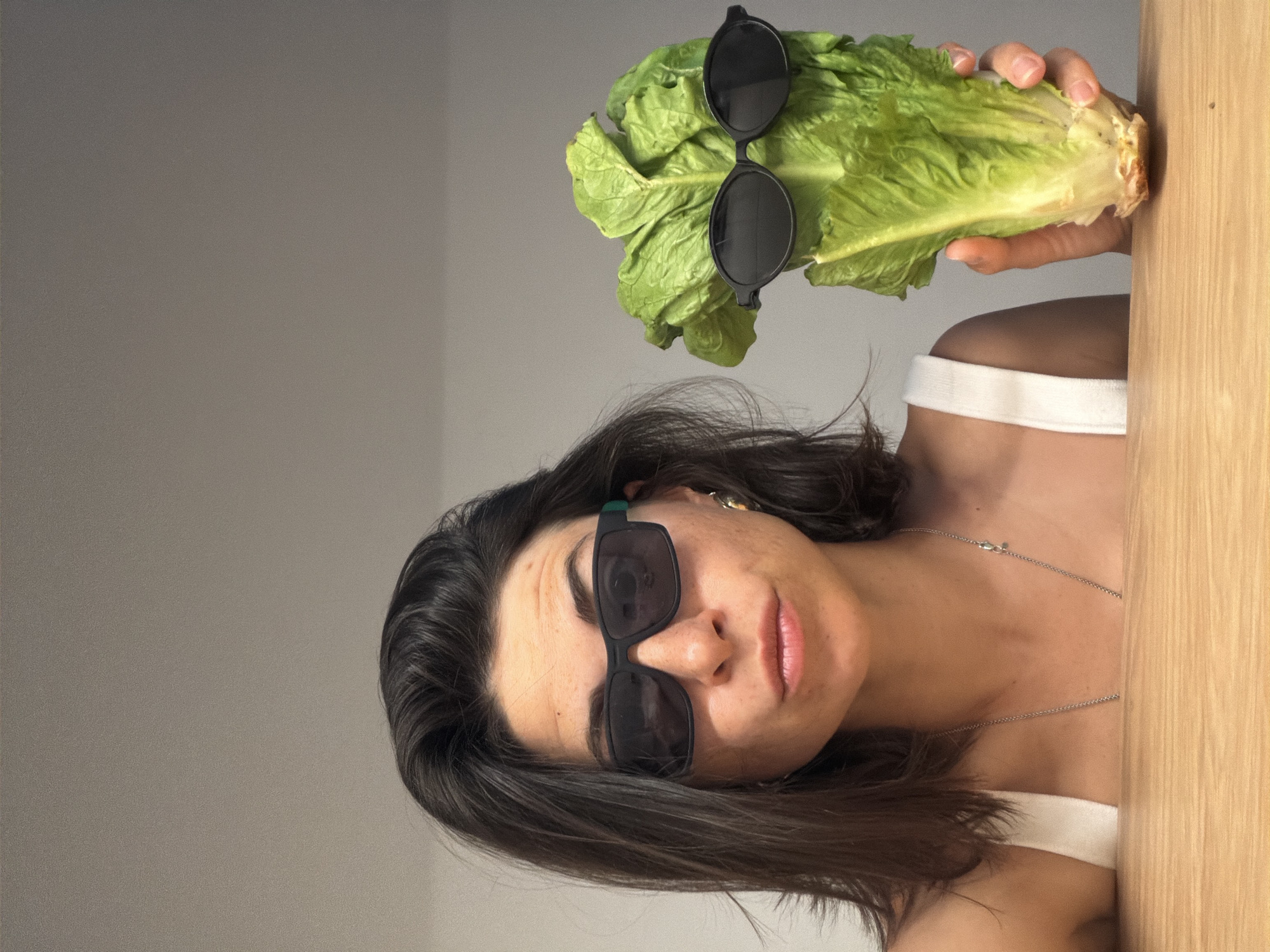 A light-skinned woman with brown hair and wearing dark sunglasses poses with a head of romaine lettuce, which is also adorned with dark sunglasses. She has a silver necklace and wears a white top. The background consists of a grey wall and a wooden door. This image's location cannot be confirmed as Budva, Montenegro.