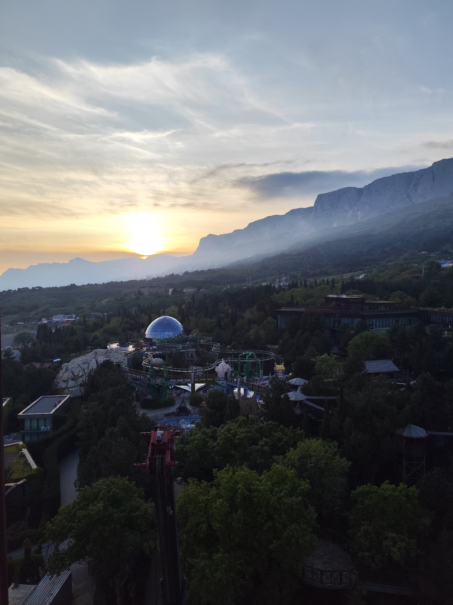 The image depicts a landscape with a setting sun and a view over a cityscape, possibly in Симеизский поселковый совет, Ukraine. The scene includes mountains in the background, partially obscured by mist or low clouds. The sun is setting on the left side of the image, casting a warm glow across the sky and landscape. In the middle ground, there is a complex of buildings, potentially including a park or entertainment area, with a prominent blue dome-shaped structure. There are also various green trees and other vegetation, suggesting a forested or park-like environment. A ride with a red metal frame is shown in the foreground. The lighting suggests it is either early morning or late evening. The overall atmosphere is serene, with the mist and soft light creating a hazy and somewhat mysterious ambiance.