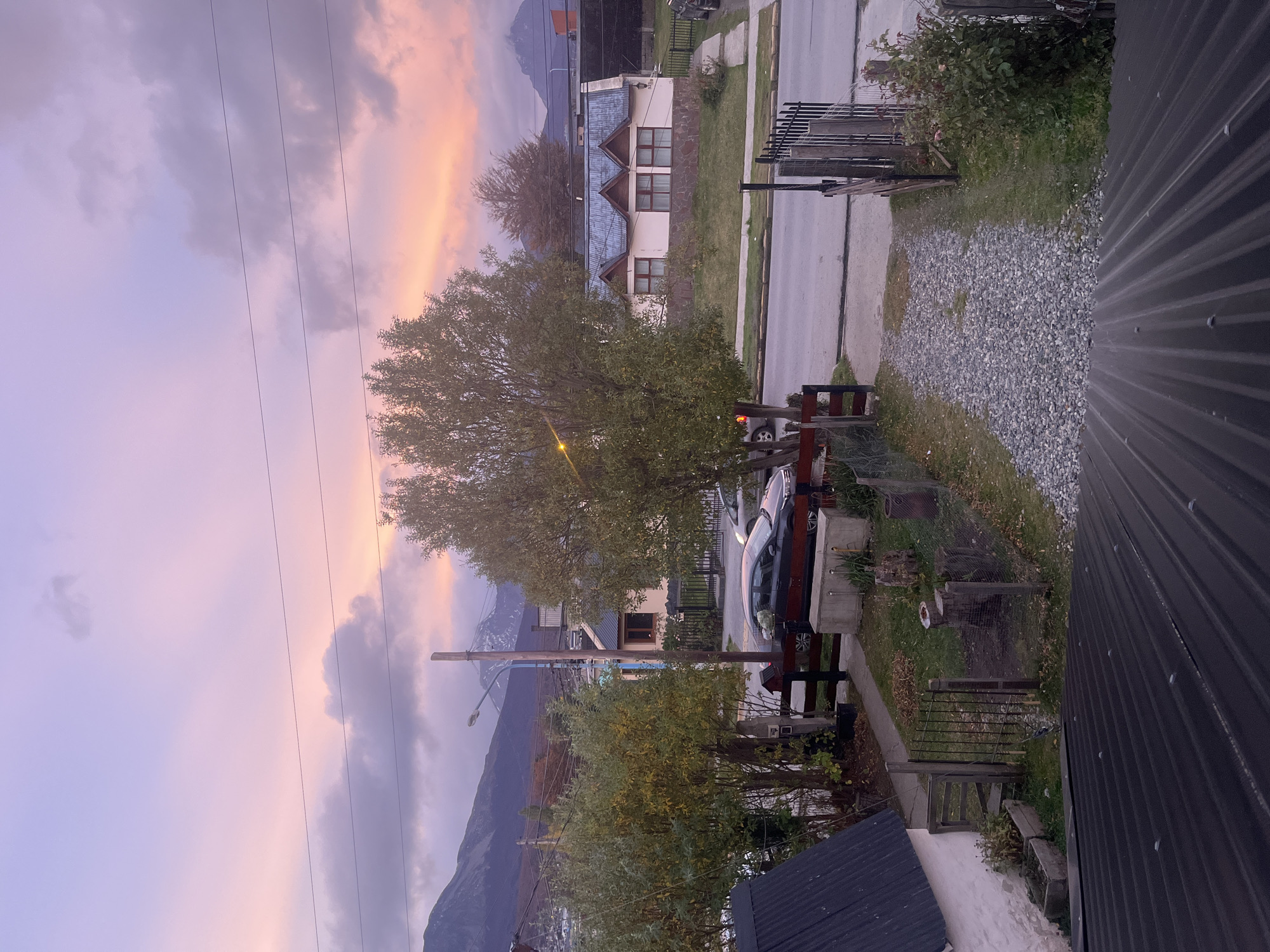 This is an eye-level, outdoor shot taken in Ushuaia, Argentina. The scene is at dusk or dawn, with a sky of pastel pink and gray clouds over a residential street. A mountain range is visible in the background. In the foreground is a corrugated metal roof. The street is lined with houses, trees, and parked cars. A streetlight is illuminated. Power lines run across the sky. The overall atmosphere is calm and peaceful.
