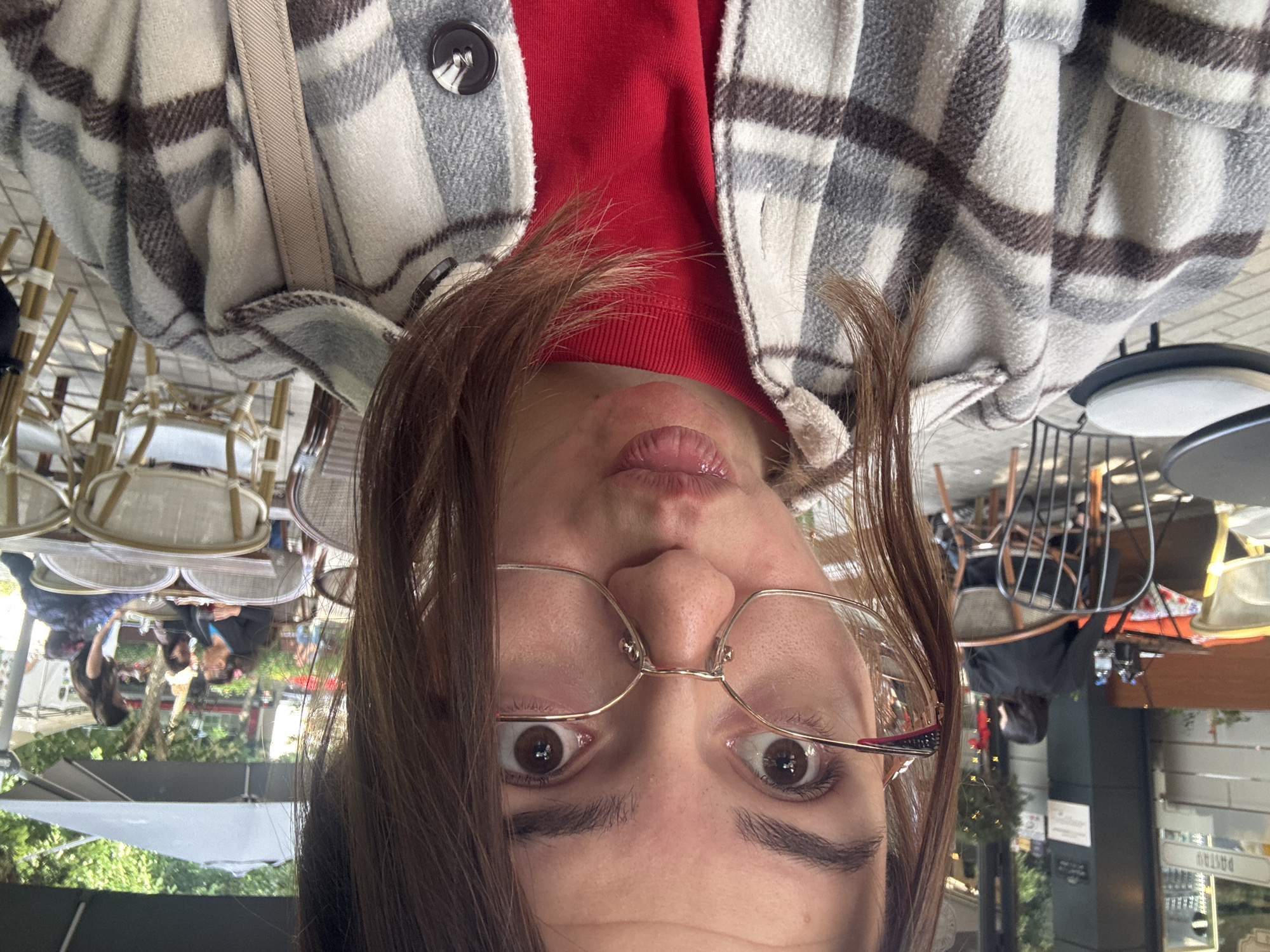 The image shows a person with brown hair and glasses, wearing a red shirt and a plaid jacket. The person is making a face with their lips puckered. The background appears to be an outdoor cafe or restaurant in an urban setting. There are tables and chairs visible, and greenery can be seen in the reflection of a window or glass surface. People are seated at the tables in the reflection. There is a building with signage visible in the reflection.