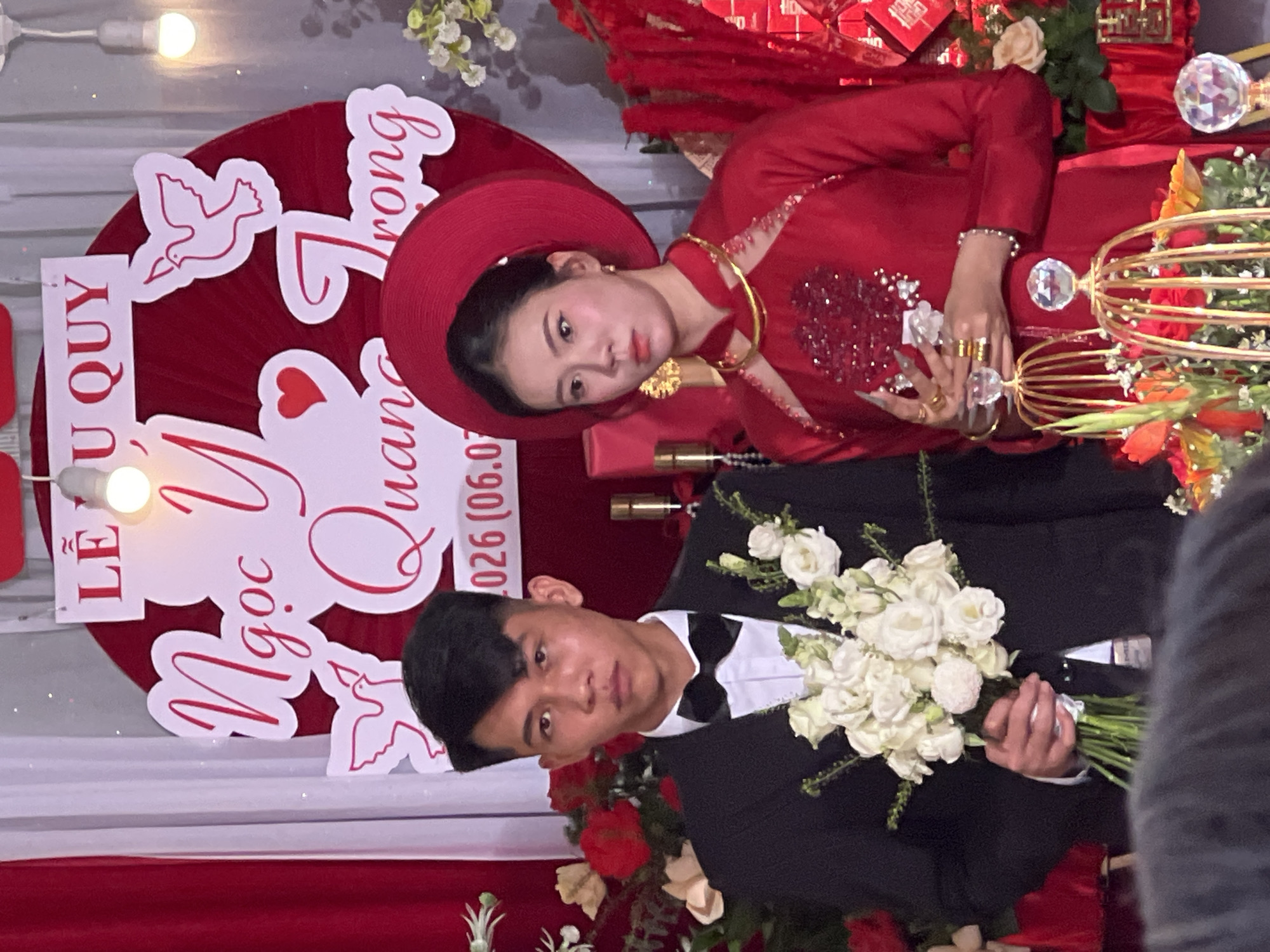 A photo shows a wedding scene with a couple in formal attire in Tay Son Commune, Vietnam. The bride wears a traditional red dress with a matching headdress and gold jewelry. She has long fingernails with decorative elements and multiple rings. The groom is in a black suit with a bow tie, holding a bouquet of white roses. Behind them is a red backdrop with white lettering, including the names "Lễ Vu Quy Ngọc Ý Quang Trong" and the date "026 (06.07)". There are floral arrangements and other wedding decorations.