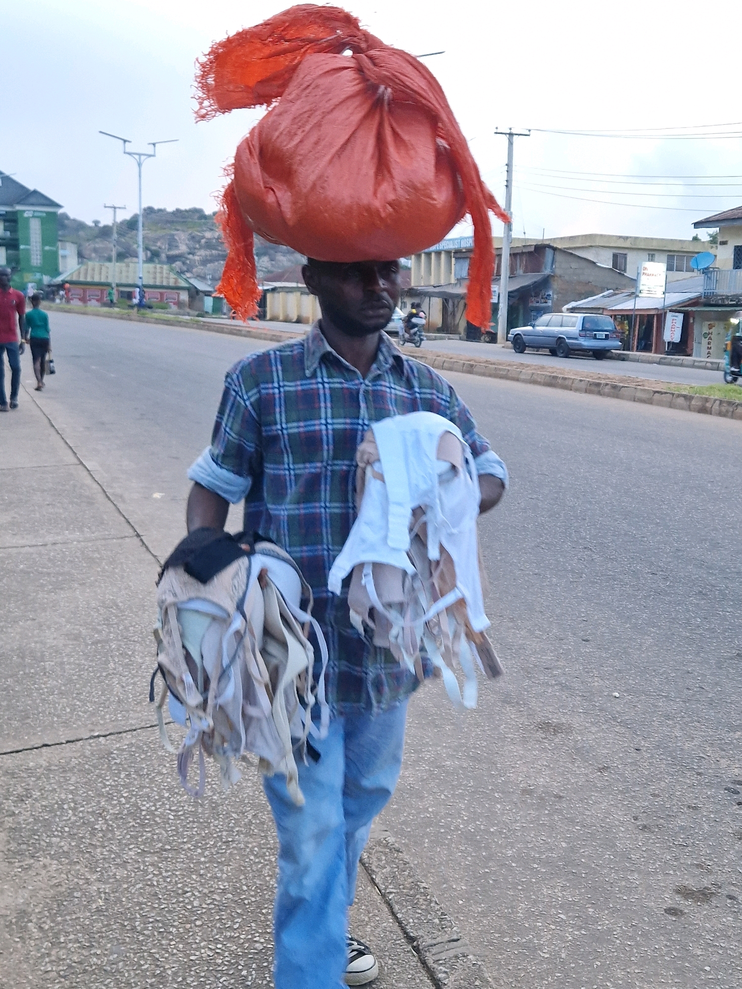 A man is walking on a paved road in Kabong, Nigeria. He is carrying a large orange bundle on his head and a pile of what appears to be used bras in his arms. The man is wearing a plaid shirt and blue jeans. In the background, there are buildings, a car, and other pedestrians. The sky is overcast.