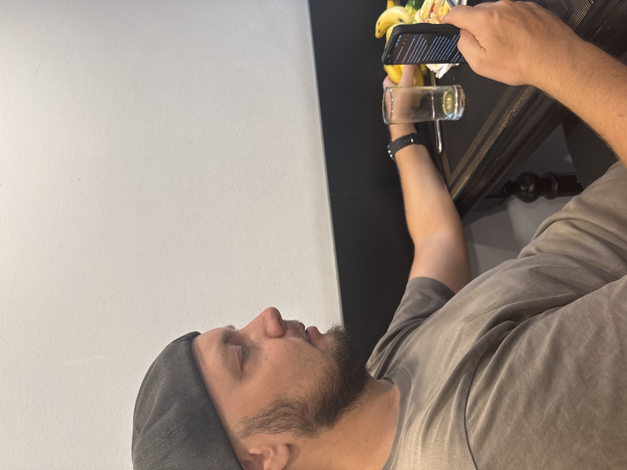 A man with a beard and a grey hat is taking a photograph of a drink and food on a black surface using a smartphone. The drink is in a clear glass. The food appears to include bananas. The man is wearing a grey t-shirt and a black smartwatch. The background includes a white wall. The location cannot be determined from the image, but it is stated that it is in Hà Nội, Vietnam.