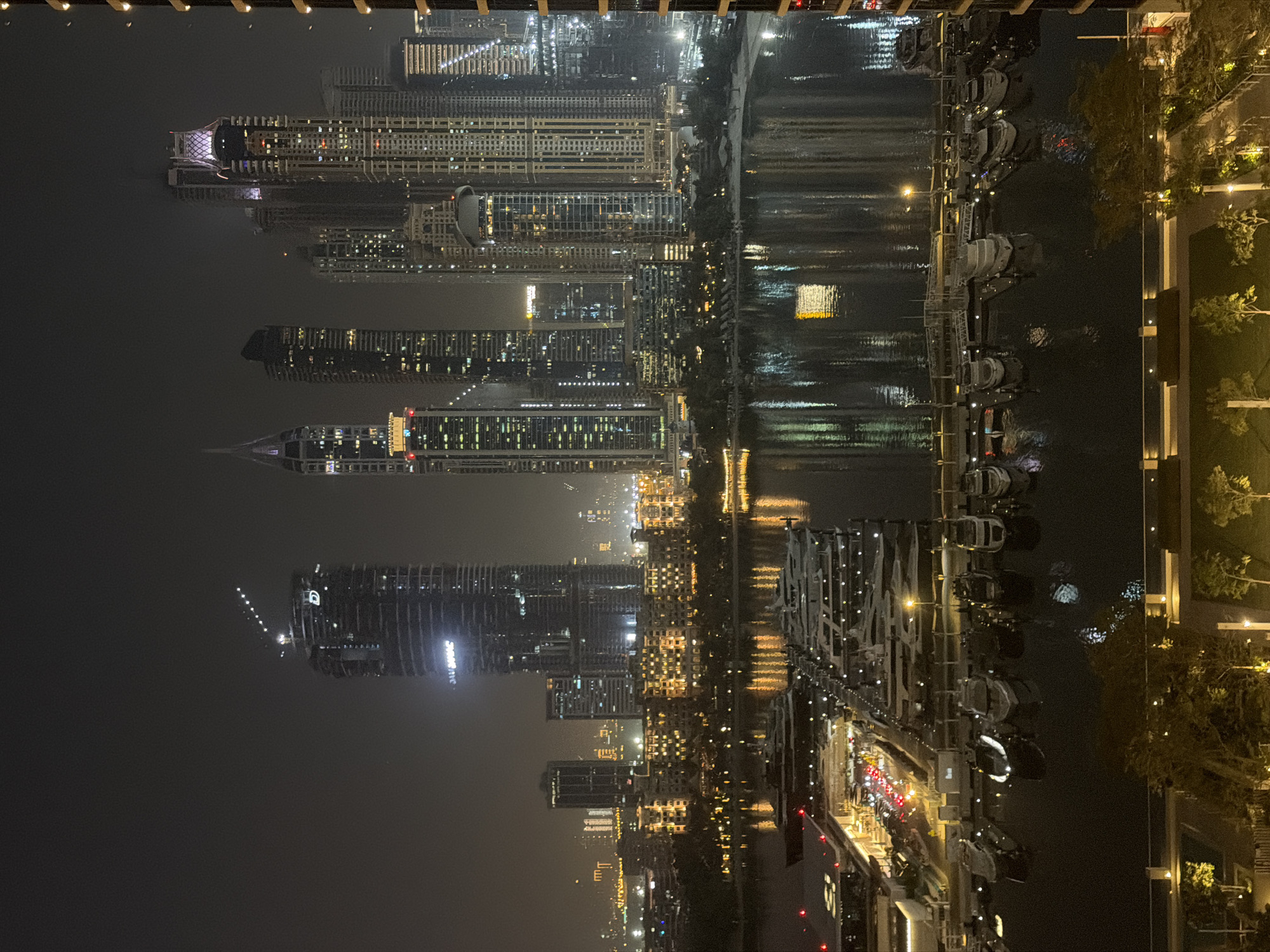 This is a nighttime aerial photograph of the Dubai Marina in Dubai, United Arab Emirates. The scene is dominated by a dense cluster of modern skyscrapers, illuminated by numerous windows and exterior lights. A wide body of water, the marina, occupies the foreground, filled with a large number of yachts docked in neat rows. The reflections of the city lights and the yachts shimmer on the dark water's surface. A promenade or walkway runs along the edge of the marina, lined with trees and lights. The overall impression is of a vibrant and affluent urban landscape at night.