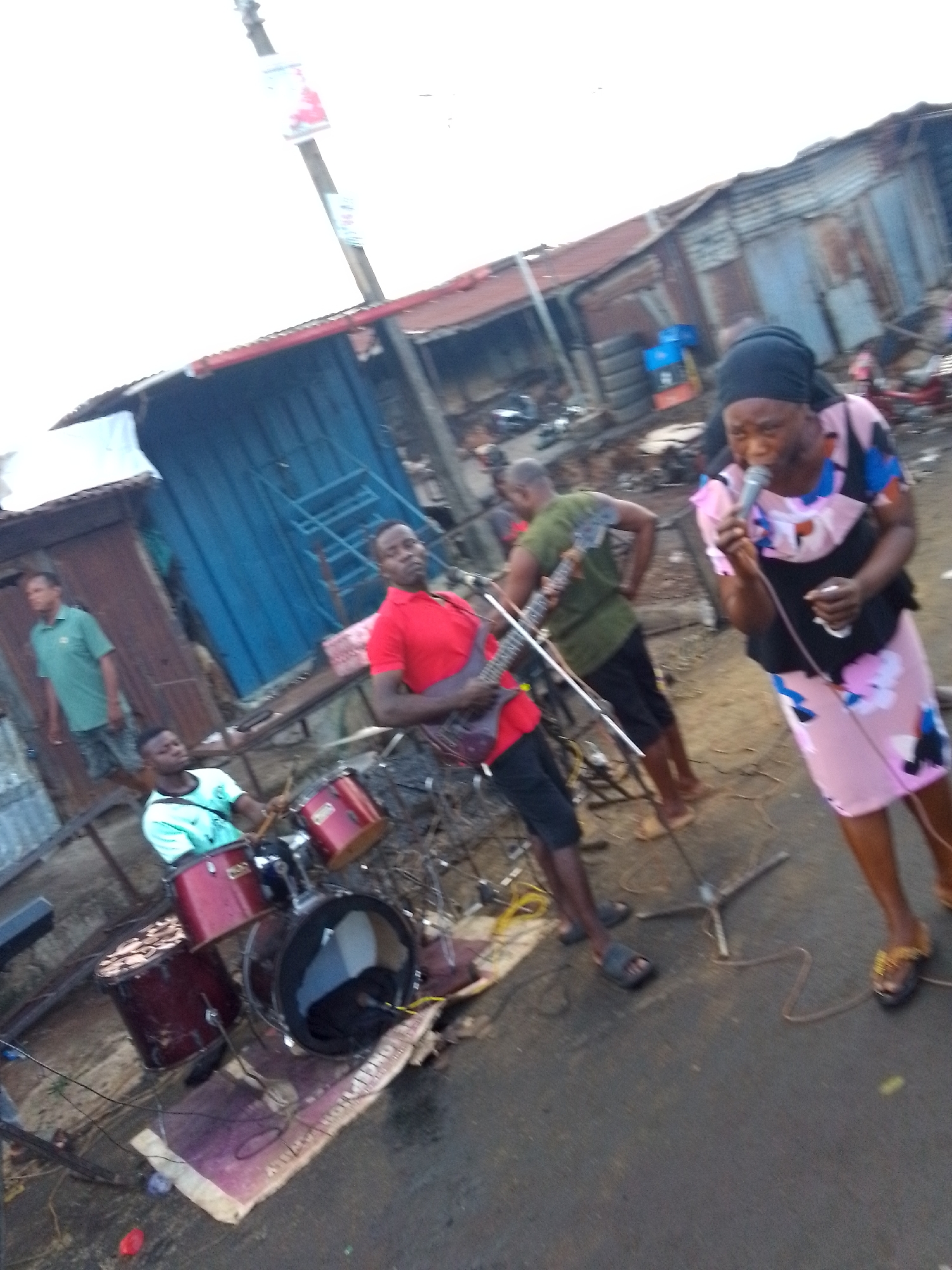 The image depicts a street performance by a band in Onitsha, Nigeria. The band consists of a drummer, a guitarist, and a female vocalist who is singing into a microphone. The drummer is positioned behind a small red drum set. The guitarist, wearing a red shirt, is playing his instrument. The vocalist, wearing a floral dress and a headwrap, is holding a microphone. The band is surrounded by electrical wires.
In the background, there are several people watching the performance. Several corrugated metal structures are visible, along with tires, other discarded materials, and a blue staircase. A utility pole with a banner is on the left. The setting suggests an improvised, informal stage area in a busy street.