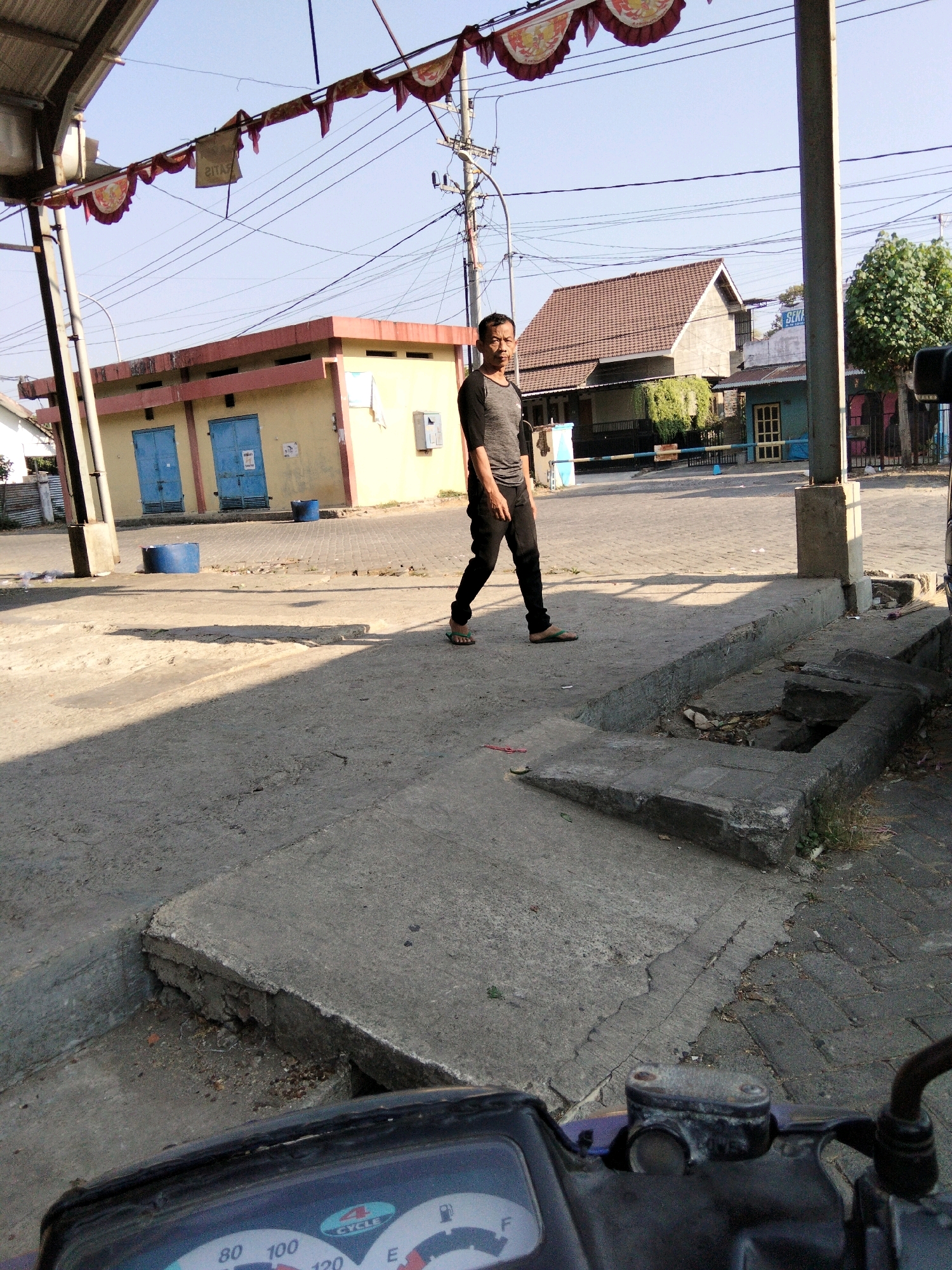 The image depicts an outdoor scene in what appears to be a semi-urban or rural area, possibly in Banyudono, Indonesia. In the foreground, the handlebars and instrument panel of a scooter are visible, suggesting the photo was taken from the perspective of someone on the scooter. A man in a dark grey t-shirt and black pants is walking across a concrete path in the mid-ground. He is looking towards the camera. Behind him, there is a light yellow building with two blue doors. Overhead, there are power lines and decorative banners. In the background, residential buildings with tiled roofs and trees can be seen. The ground is a mix of concrete and pavers. The scene is illuminated by natural daylight.