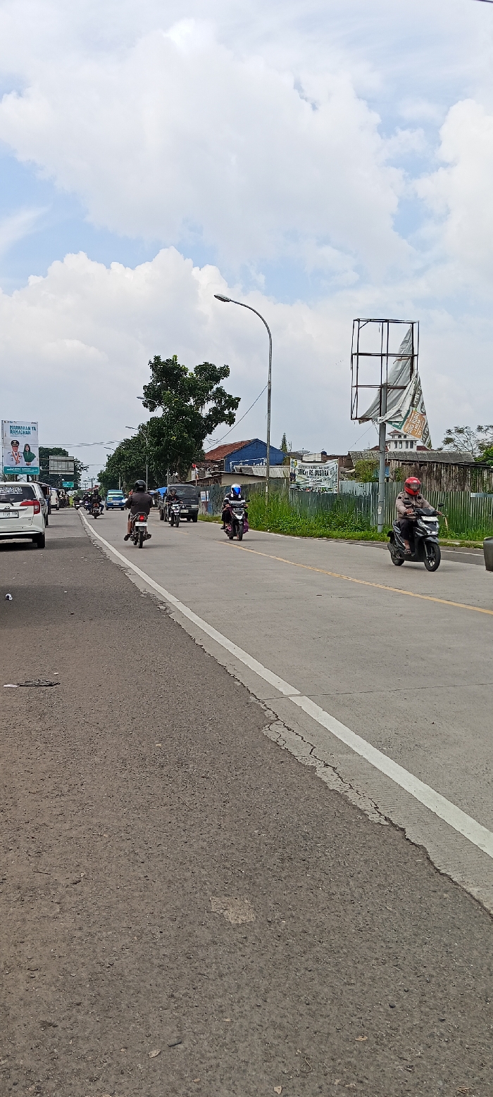 The image shows a street scene in Kutawaringin, Indonesia, taken at latitude: -6.994, longitude: 107.532. The street is paved and lined with a single white line down the center. The street has a few cracks in the asphalt, especially along the edges. There are six motorcycles traveling down the street. Four of the motorcycles are black and white, and two are black and red. The riders are wearing helmets. There is a light pole with a street lamp on the right side of the road. The left side of the street has a billboard with a person wearing a uniform and a woman wearing a hijab. There are several trees and bushes on both sides of the street, and a few buildings in the distance. There is a blue sky with white clouds in the background.