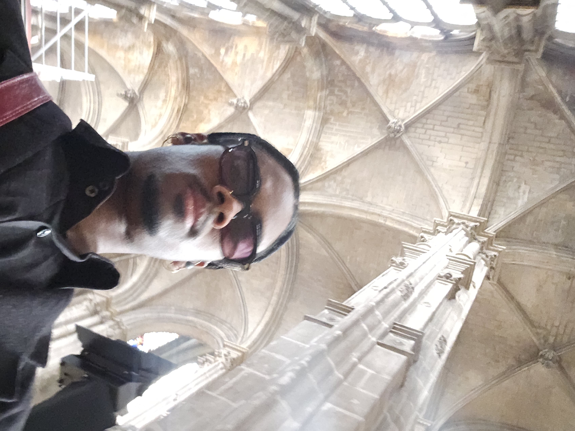 A person with dark skin and sunglasses is in the foreground, oriented so the camera is angled upward. The person is wearing a black jacket with a red trim on the shoulder. The background features the interior of a grand building with stone columns and an elaborately decorated ceiling. The ceiling has a repeating pattern of arched sections converging at decorative points. Light is filtering in from above through windows, which suggests the scene is taking place during daylight hours. The image is angled, so the person and the architecture are viewed from a low perspective. It cannot be determined if this image is from Paris or elsewhere.