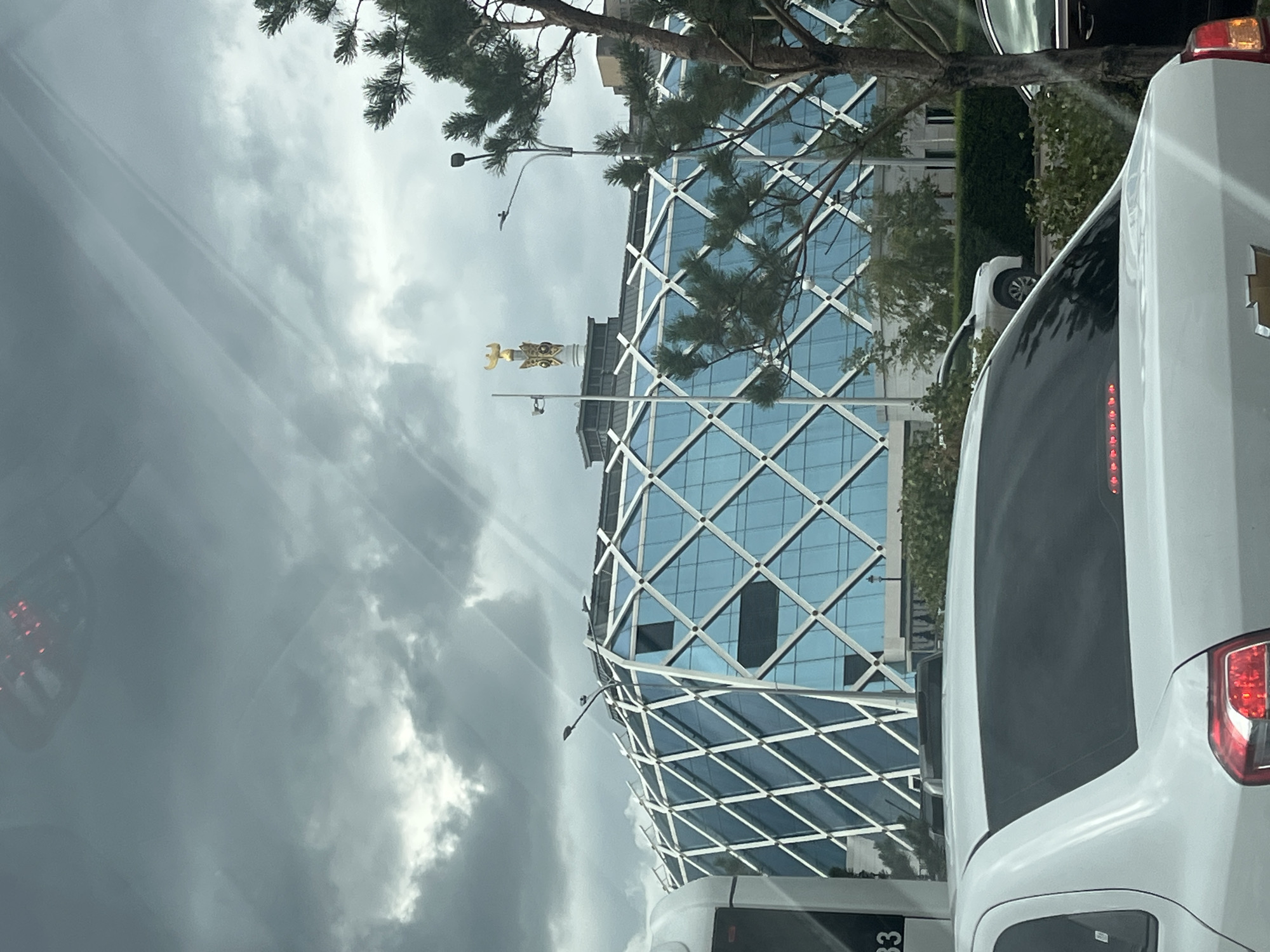 A modern, glass-facaded building with a diamond-shaped lattice structure is visible in Astana, Kazakhstan, under a cloudy sky. The building is topped with a golden sculpture. In the foreground, the back of a white car is visible, obscuring the lower part of the building. Trees and other vehicles are also present in the scene.