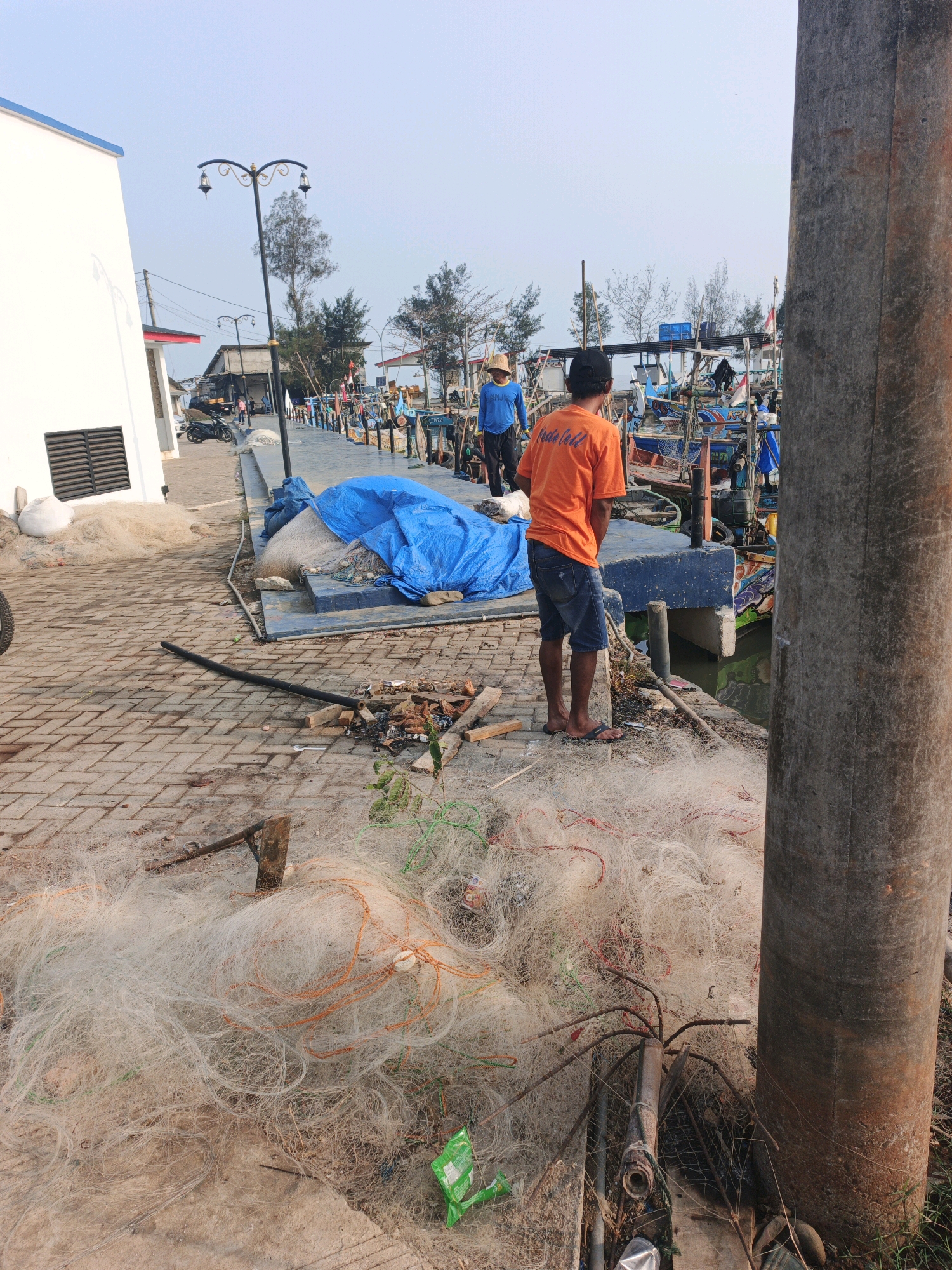In Unknown, Indonesia, a concrete pier, cluttered with fishing nets and debris, leads to a body of water where several colorful fishing boats are docked. A large, rough-textured, cylindrical pillar stands prominently on the right side of the frame. On the pier, two men are visible. One, wearing an orange t-shirt and denim shorts, stands with his back to the camera, looking towards the boats. Another man, dressed in a blue t-shirt and dark pants, walks away from the camera on the pier. A white building with a blue trim is situated on the left side of the image, adjacent to the pier. The sky above is clear and light.