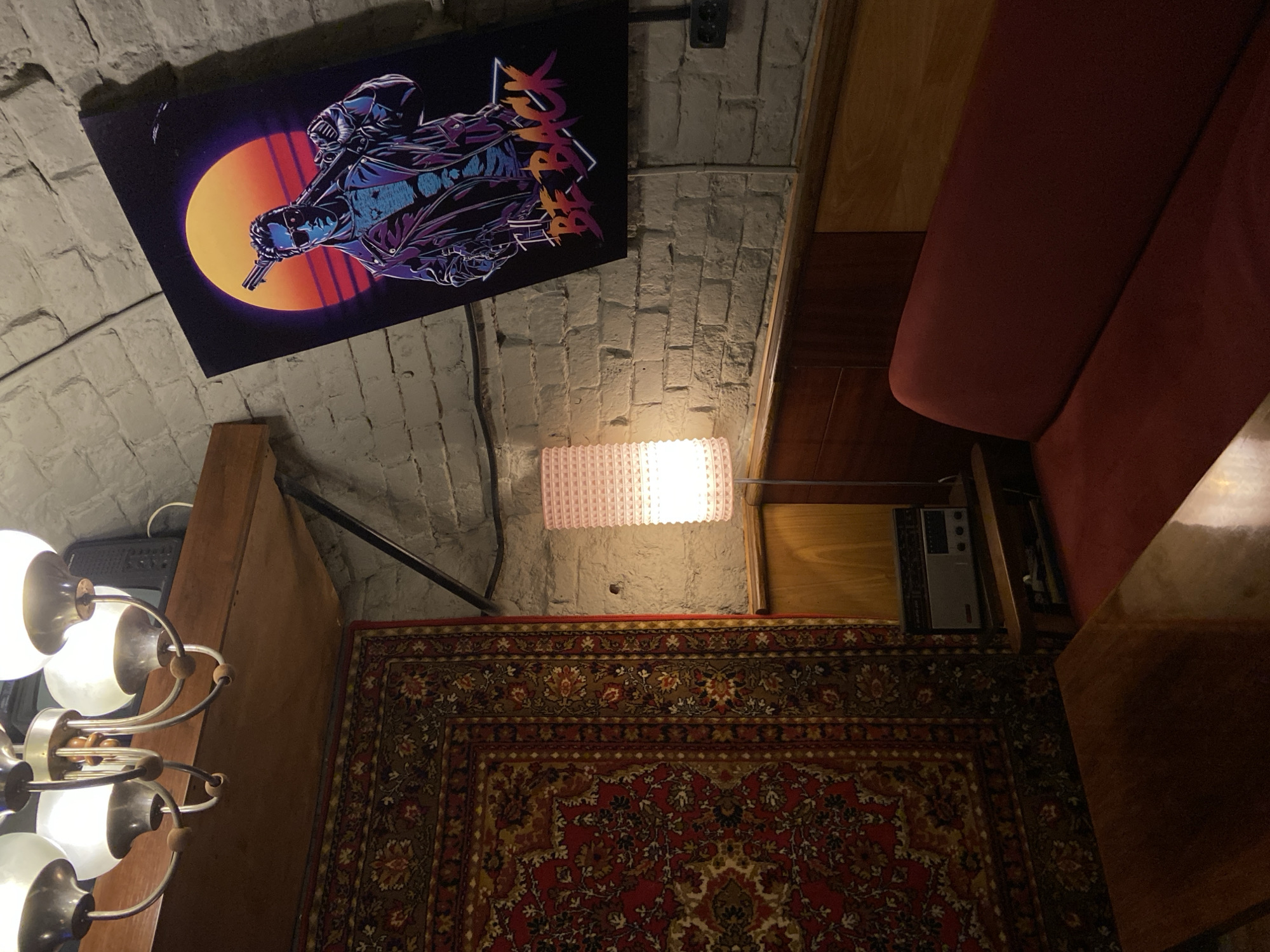 The image shows an indoor scene with a mix of vintage and modern elements. On the ceiling, there is a poster featuring a character against a sunset backdrop, mounted on a white brick wall. Beneath the poster, a small, textured lamp emits a warm light. A corner of a red upholstered bench or sofa is visible along the right side. Below, a patterned rug covers the floor, and next to it, a vintage-style radio or appliance sits on the rug. A chandelier with multiple lights is partially visible on the left. The location is not specified, and no people are visible.