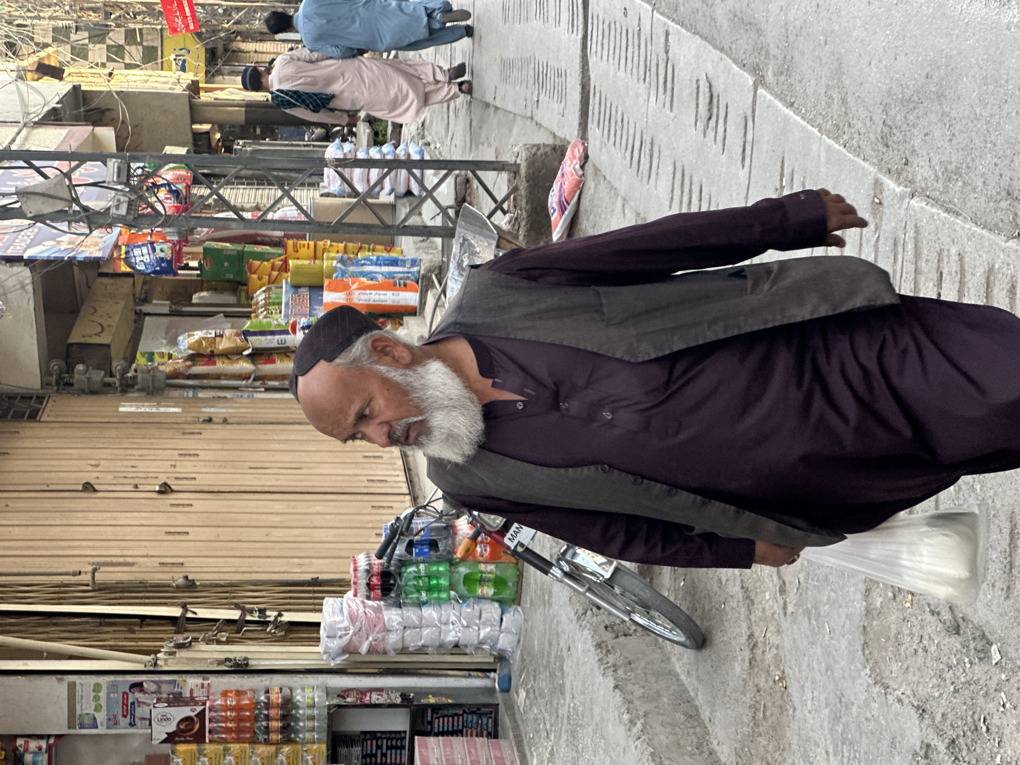 In Quetta City Tehsil, Pakistan, an elderly man with a white beard, wearing a black cap, dark purple button-down shirt, and grey vest, walks down a concrete sidewalk. He holds a plastic bag filled with white items in his right hand. Next to him is a bicycle laden with supplies, including stacks of bottled drinks and packages of goods. Behind him is a store with a closed metal roll-up door, displaying various products on shelves. In the background, other people can be seen walking along the sidewalk near additional shops. Overhead, a network of wires and supports crisscrosses the area.