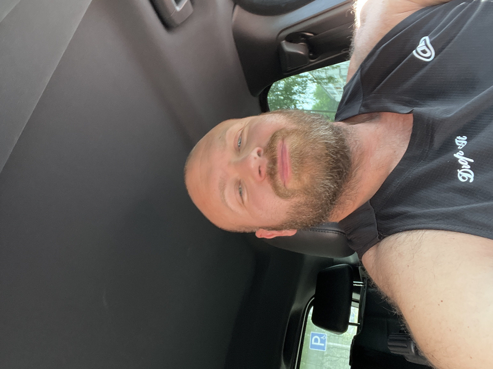 A bald, light-skinned man with a beard is seated in the driver's seat of a vehicle. He is wearing a black tank top with white lettering that says "Drop it" and a logo on the right side. The interior of the car is visible, including the dashboard and headliner. Through the window, a blue and white sign with a 'P' and a wheelchair symbol is visible, indicating a parking spot. The background outside the window shows trees and buildings, suggesting an urban or suburban setting. The man's face is turned towards the camera, and he appears to be smiling slightly. There is no indication of the specific location in the city of Noviy, Russia within the image itself.