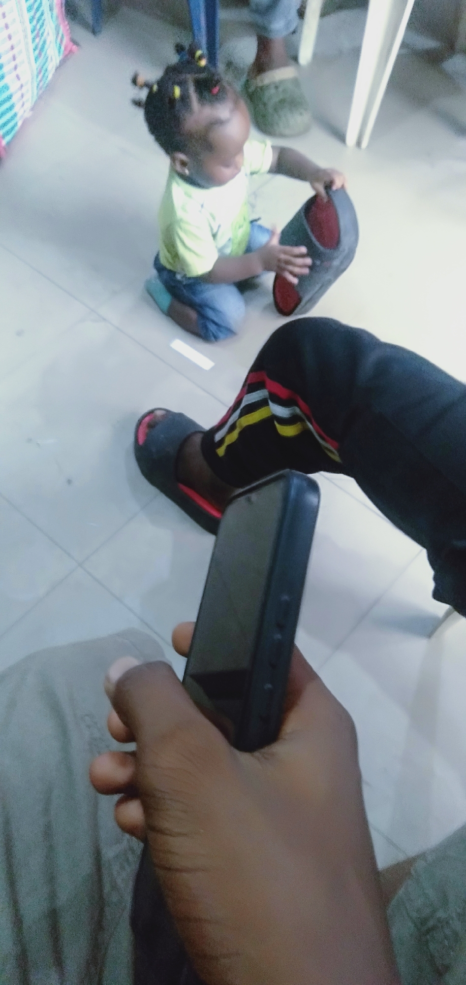 In Shangisha, Nigeria, a child with braided hair and colorful beads is kneeling on a tiled floor, wearing a light green t-shirt and blue pants. The child is holding a dark-colored shoe with a red interior. To the right, an adult's legs are visible, wearing black pants with red, yellow, and white stripes and dark slides. The adult is holding a dark smartphone. The environment appears to be indoors with tiled flooring and furniture in the background, suggesting a residential or public indoor space.