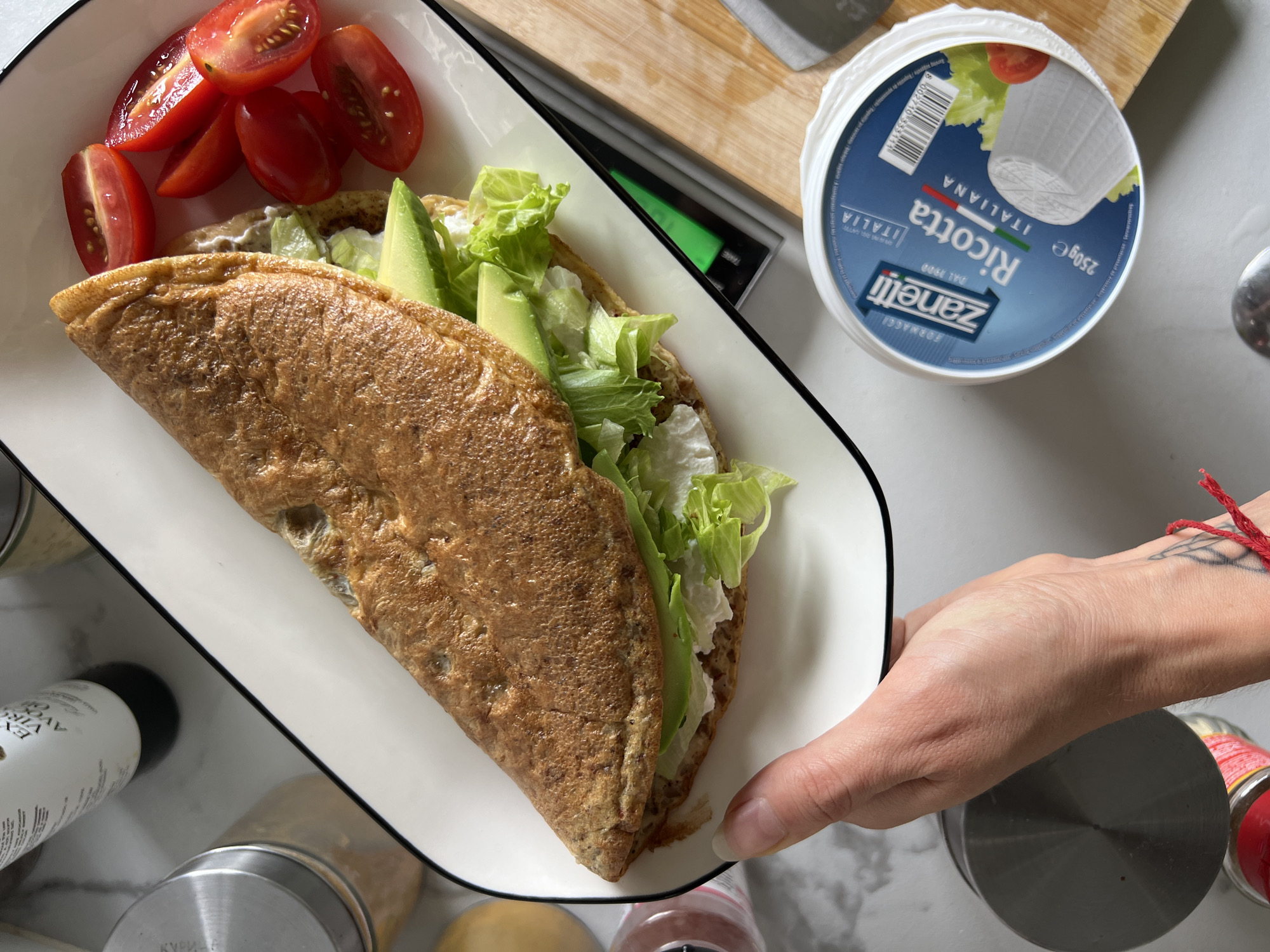 Here is a detailed description of the image:
The image shows a white rectangular plate with a black rim held at an angle by a person's hand. The plate contains a folded crepe or omelet filled with lettuce and avocado slices, accompanied by halved cherry tomatoes on one side.
To the right of the plate, there's a container of "Zanetti Ricotta Italiana" cheese. Partially visible behind the cheese is a cutting board with a knife. Several spice containers are situated around the plate on what appears to be a marble countertop. The hand holding the plate has a bracelet and a tattoo on the wrist.