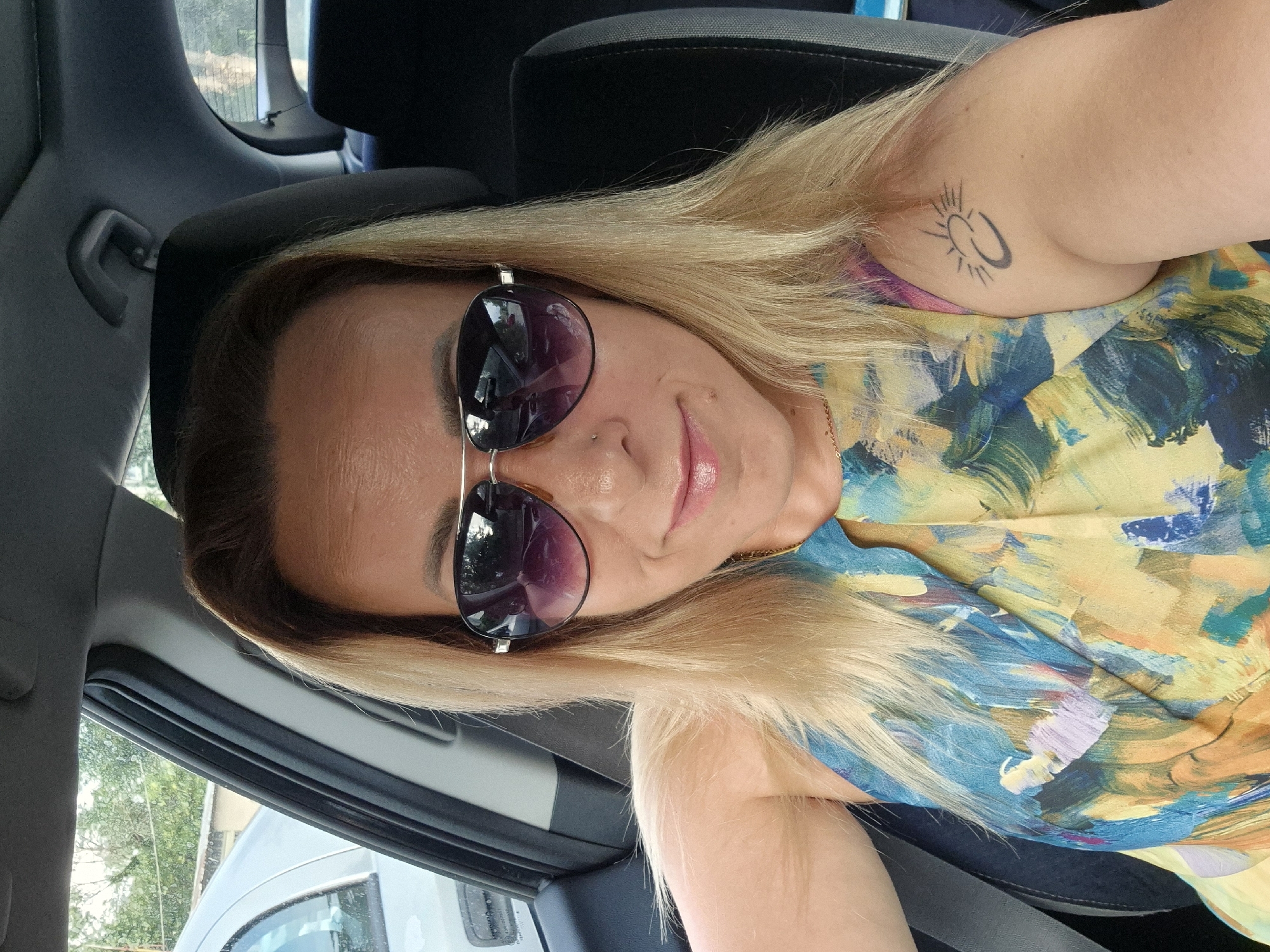A blonde woman wearing sunglasses and a colorful sleeveless dress is taking a selfie inside a car. She has a small tattoo of a sun on her left arm. The car appears to be parked, with other vehicles and trees visible through the windows. The lighting suggests it is daytime. The overall scene is casual and personal.