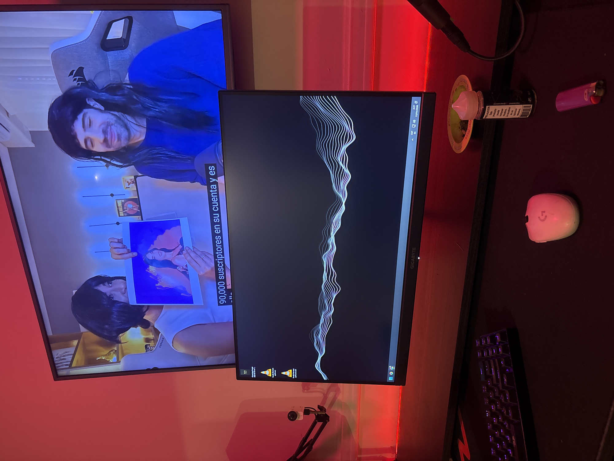 The scene depicts a desk setup with a television displaying a video of two individuals, one holding a picture of two women, with the text "90,000 suscriptores en su cuenta y es" overlaid. The person in the video has a wig on. Beside the television is a computer monitor with an abstract white wave pattern on a dark background, with icons visible in the lower left corner. A pink lighter, e-juice and a wireless mouse are to the right of the monitor, alongside part of a keyboard. The environment is illuminated with red lighting.