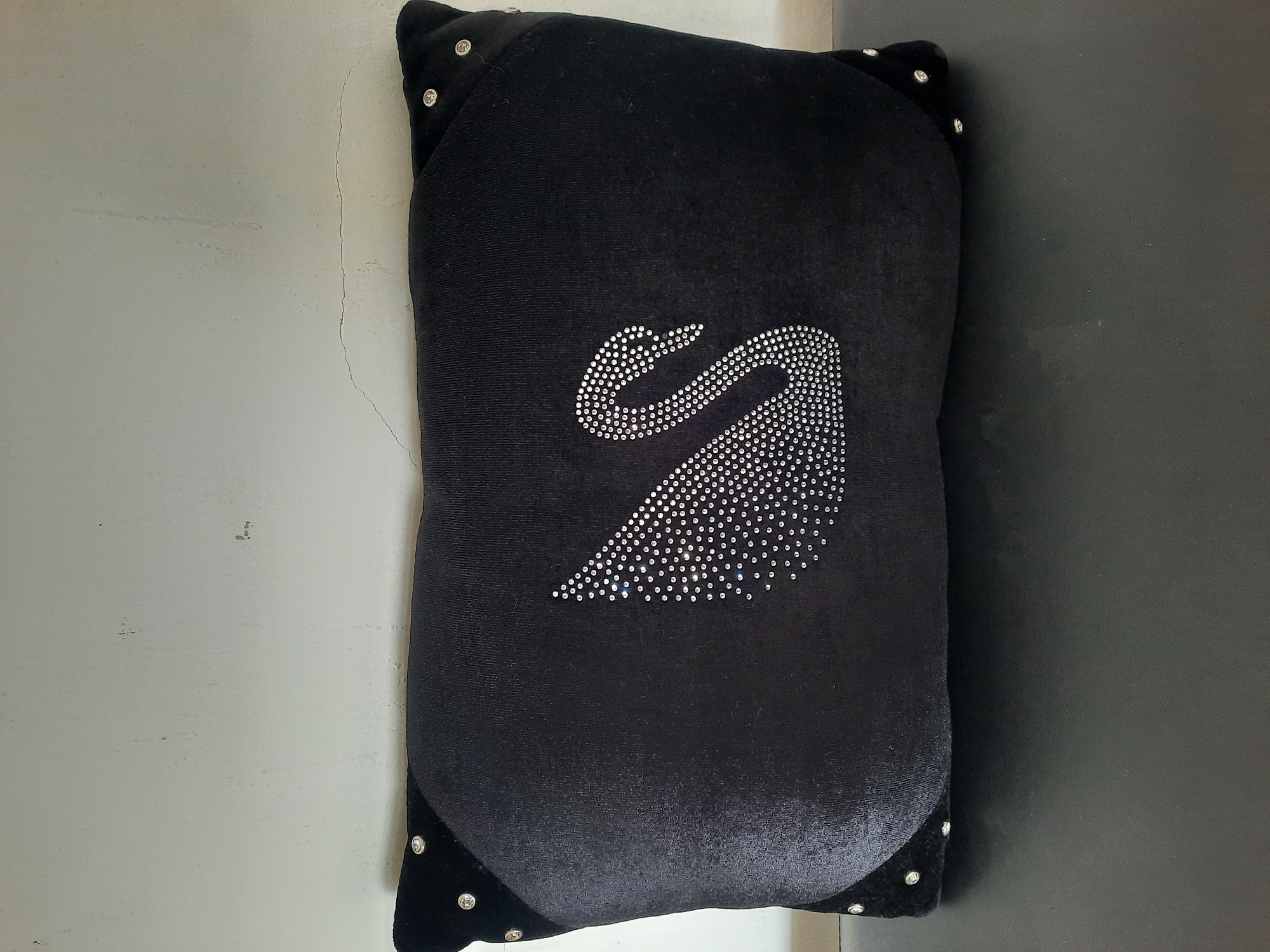 A black velvet rectangular cushion with a silver rhinestone swan design is centrally placed against a light-colored, textured wall. The cushion has black velvet corners adorned with small silver studs. The swan is positioned on the left side of the cushion, facing right. The background wall has visible imperfections and faint vertical lines. The cushion rests on a dark, flat surface. The image is a straightforward, eye-level shot of the object. The location in Lhokseumawe, Indonesia, is not discernible from the visual information provided in the image.