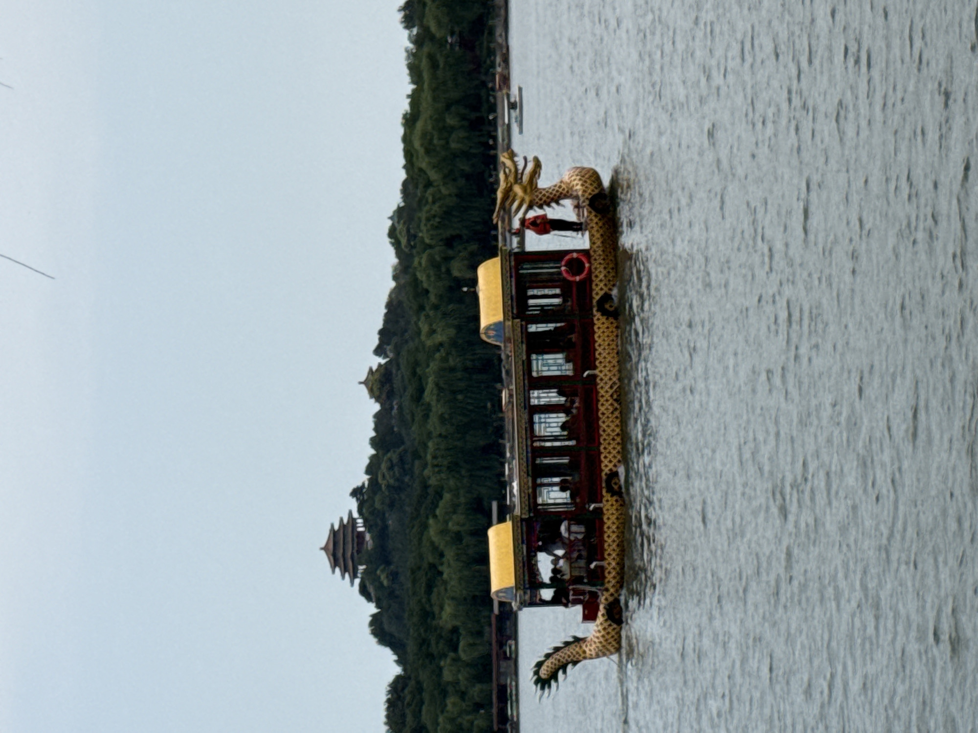 A dragon boat is sailing on a lake in front of a hill covered with trees, with a pagoda on top. The boat has a dragon head at the front and a dragon tail at the back, both made of gold. The boat is decorated with red and yellow colors. There are people inside the boat. The sky is overcast, suggesting it might be a cloudy day. The image is taken from a landscape perspective. The location is Xicheng District, China.
