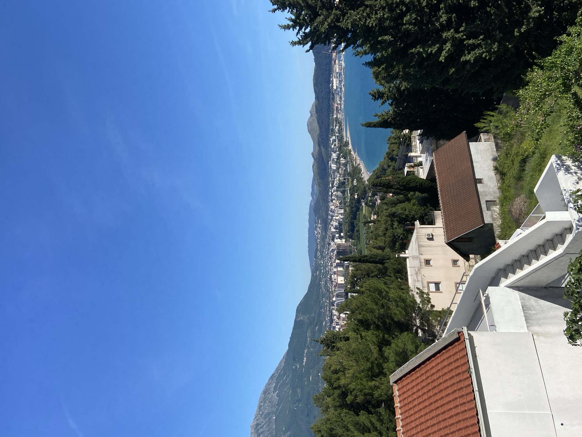 Captured at a high angle, the scene displays a coastal landscape featuring Šušanj, Montenegro. The Adriatic Sea stretches along the coastline, flanked by a strip of urban development with buildings densely packed together. Mountainous terrain forms a backdrop to the urban sprawl. Foreground elements include residential buildings with distinctive tiled roofs, interconnected by staircases leading down the hillside. Lush greenery and trees dot the landscape. Above, a clear blue sky dominates the upper portion of the frame. The image suggests a sunny day.