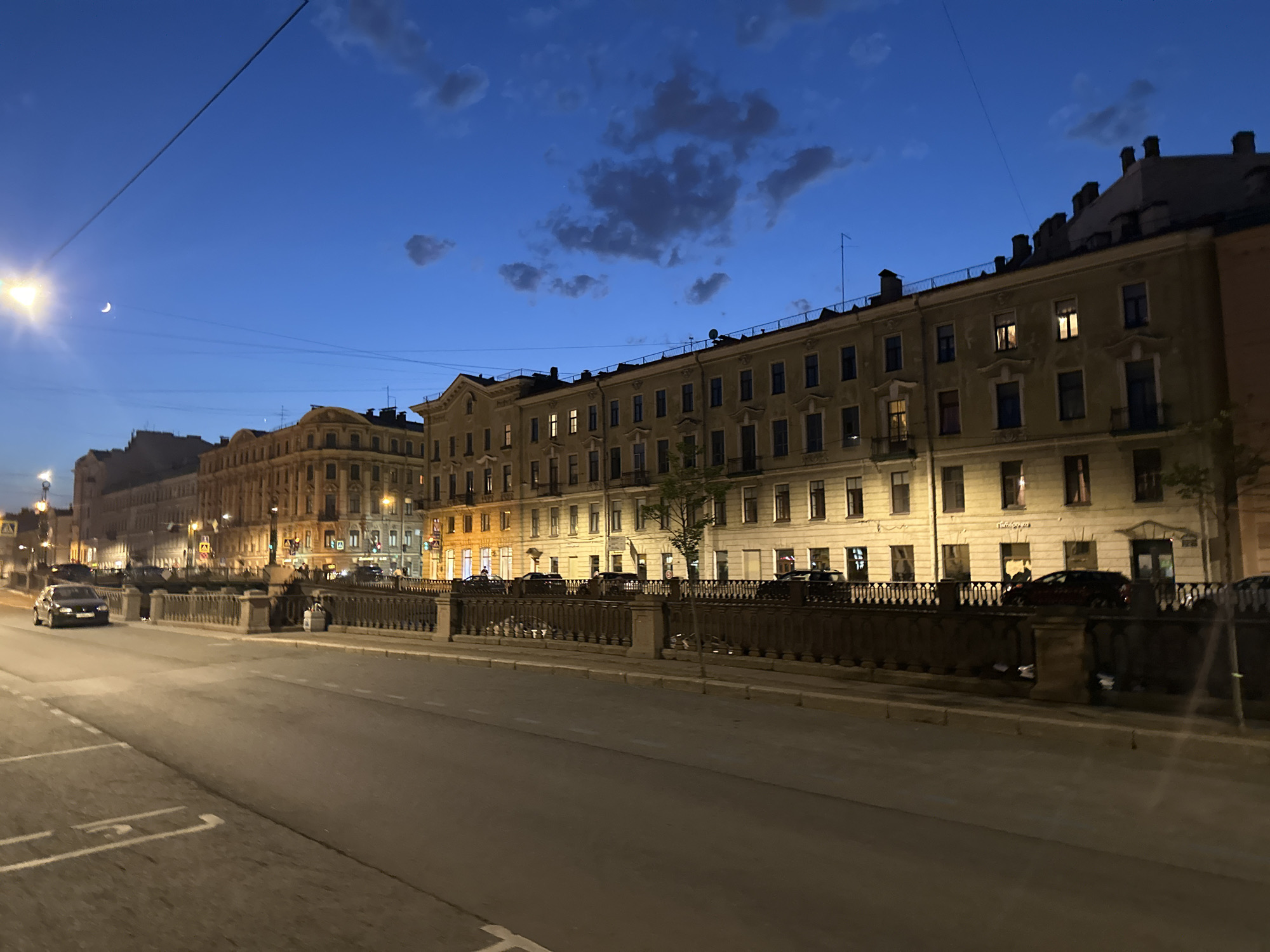 The image depicts a nighttime scene in Saint Petersburg, Russia. The primary focus is a street running parallel to a canal or river, separated by a low stone embankment with ornate metal railings. On the opposite side of the canal is a row of stately, multi-story buildings in the traditional style of the city's architecture, displaying lit windows against the backdrop of the twilight sky. Several cars are visible parked along the street adjacent to the buildings and driving on the main road. A street lamp is visible on the left of the image. The sky is a deep blue with scattered clouds. The lighting suggests the scene is captured during dusk or shortly after sunset, as the streetlights and building lights illuminate the surrounding area.