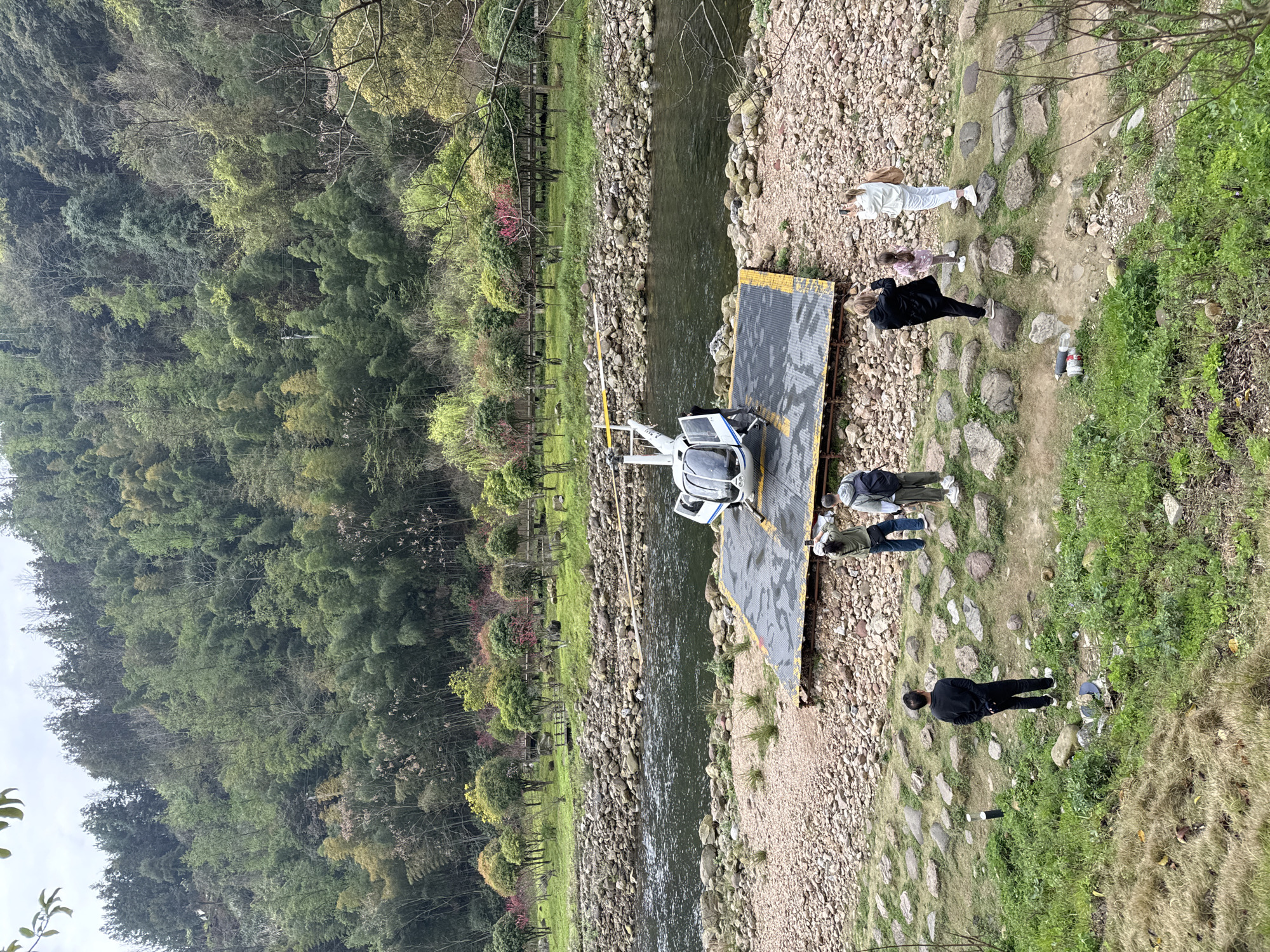 This image depicts an outdoor scene in Wulingyuan, China, featuring a parked helicopter and multiple observers by a river, framed by forested hills under an overcast sky.
A white helicopter with blue and black stripes is stationary on a dark gray, rectangular landing pad with yellow edge markings. The landing pad rests on a wide, light-colored rocky bank adjacent to a dark, flowing river. The helicopter's main rotor blades are positioned above the fuselage, and the cockpit door on the left side (from the viewer's perspective) is open. An individual, partially obscured, stands near the open helicopter door.
Eight people are visible in the scene. Three individuals are positioned on the rocky bank near the helicopter. One person, wearing an olive-green jacket, light pants, and a white hat, holds a smartphone, apparently photographing the helicopter. Another man, wearing a light gray hoodie and dark pants with a backpack, stands nearby, observing. The third individual is partially obscured near the helicopter door.
Five additional people are located on a slightly elevated, grassy path interspersed with scattered large rocks, further from the river. One man in a black long-sleeved shirt and black pants stands with his back to the viewer, hands behind his back, observing the scene. Closer to the foreground, a group of four individuals stands: two women, one in a black jacket and dark pants, and another in a white jacket and white pants, are both holding smartphones, directed towards the helicopter. Between these two women stands a young child in a pink patterned jacket and light pants. A fourth individual is partially visible within this group.
The river, characterized by dark water, flows from left to right across the midground. The opposite bank features a grassy strip with numerous small, manicured trees, some displaying pink blossoms. Behind these trees, a wooden structure or walkway is visible. The background is dominated by a steep hillside densely covered with mixed forest, including a significant presence of bamboo, under a uniformly light gray, overcast sky. Two discarded plastic bottles are present on the grassy path in the foreground.