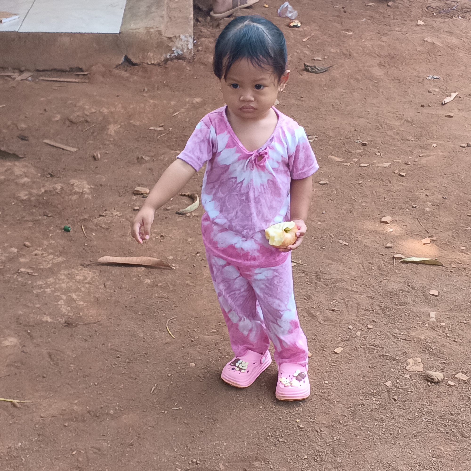 A young girl stands on a dirt path in Jayasampurna, Indonesia, holding a partially eaten apple in her right hand. She is wearing a pink and white tie-dye t-shirt and matching pants, along with pink clogs adorned with cartoon characters. The ground is a mixture of dirt, small stones, and fallen leaves. In the background, a textured concrete structure is visible. The lighting suggests it is daytime, and the overall environment appears to be outdoors, possibly in a residential or communal area.